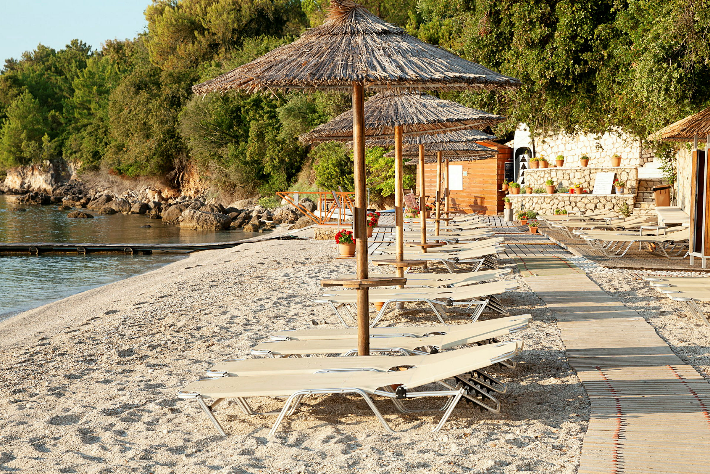 Solstolar och parasoller på en strand med en trästig och grönska i bakgrunden. (AI-genererad text)