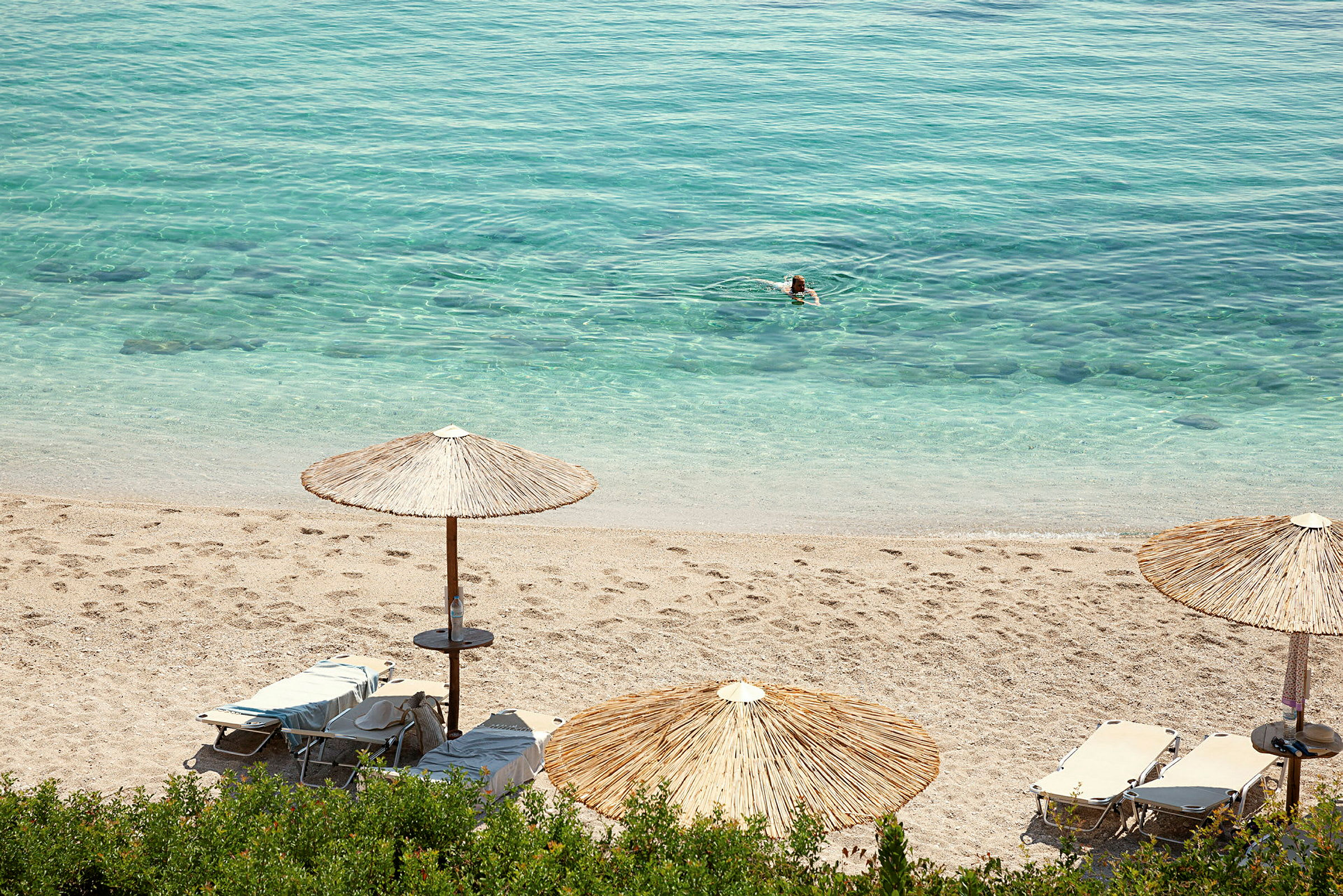Strand med parasoller och solstolar, klarblått vatten med en simmande person. (AI-genererad text)