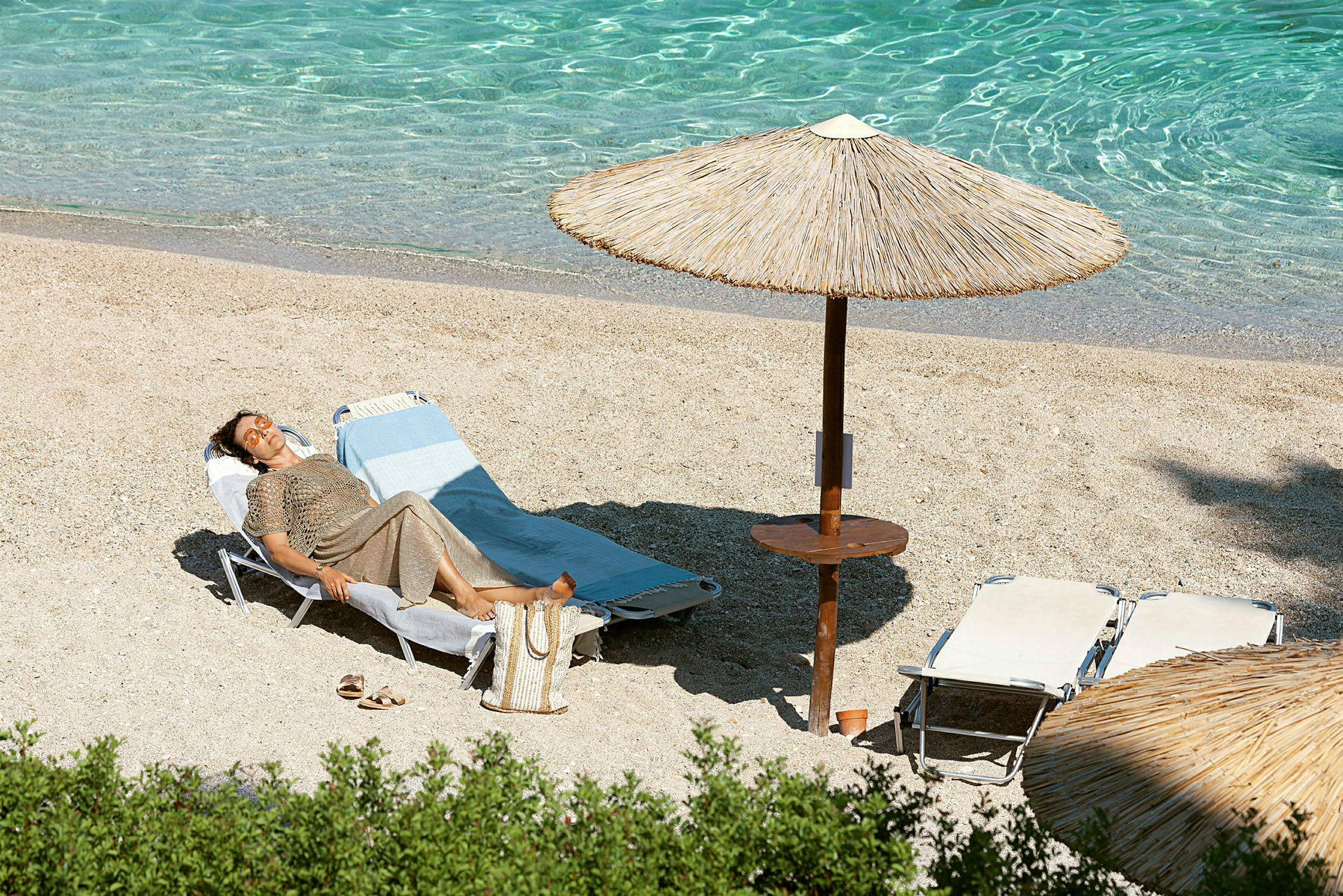 En person ligger på en solstol vid stranden under ett parasoll bredvid klart vatten. (AI-genererad text)