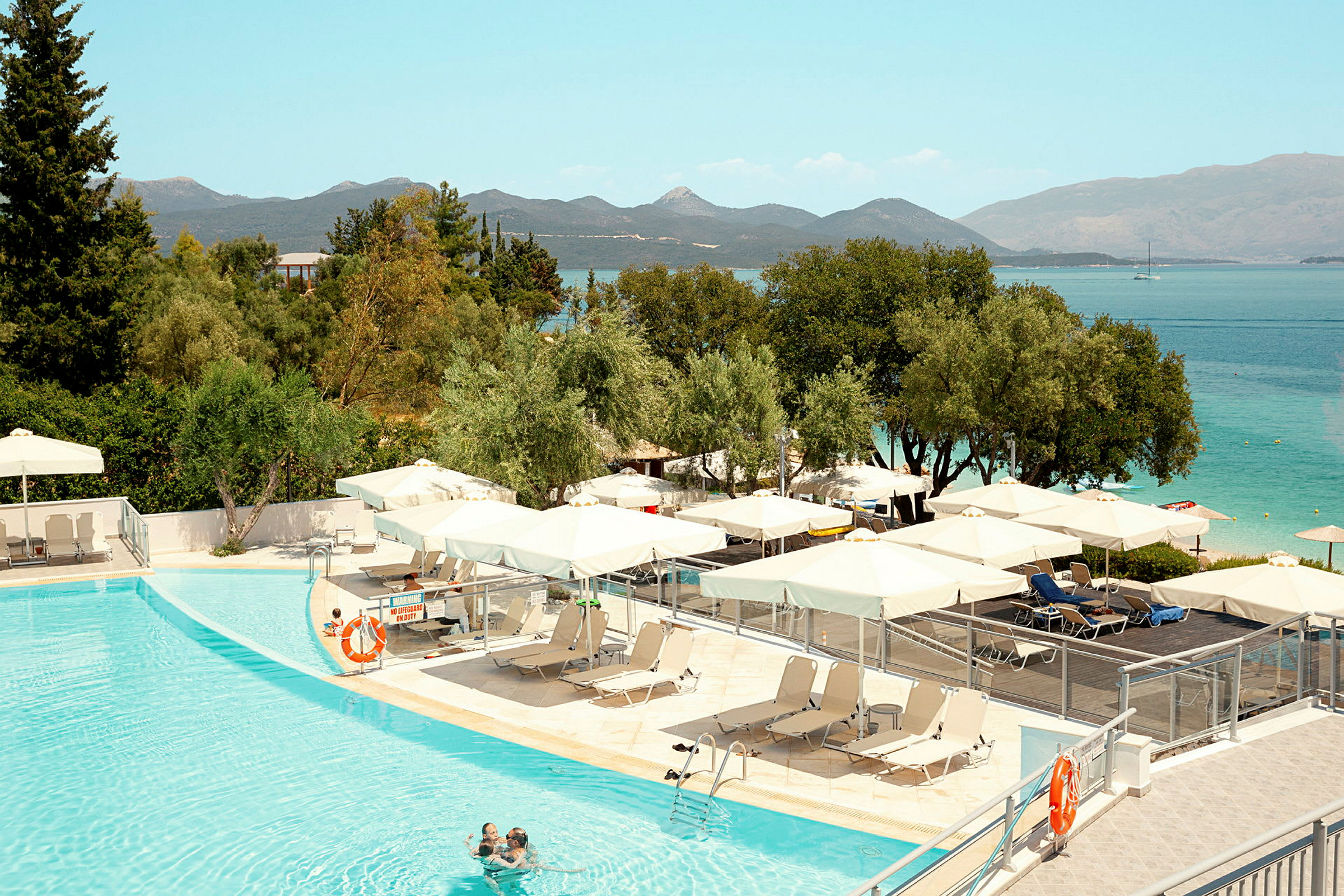 Poolområde med solstolar och parasoller, omgiven av grönska och utsikt över havet. (AI-genererad text)
