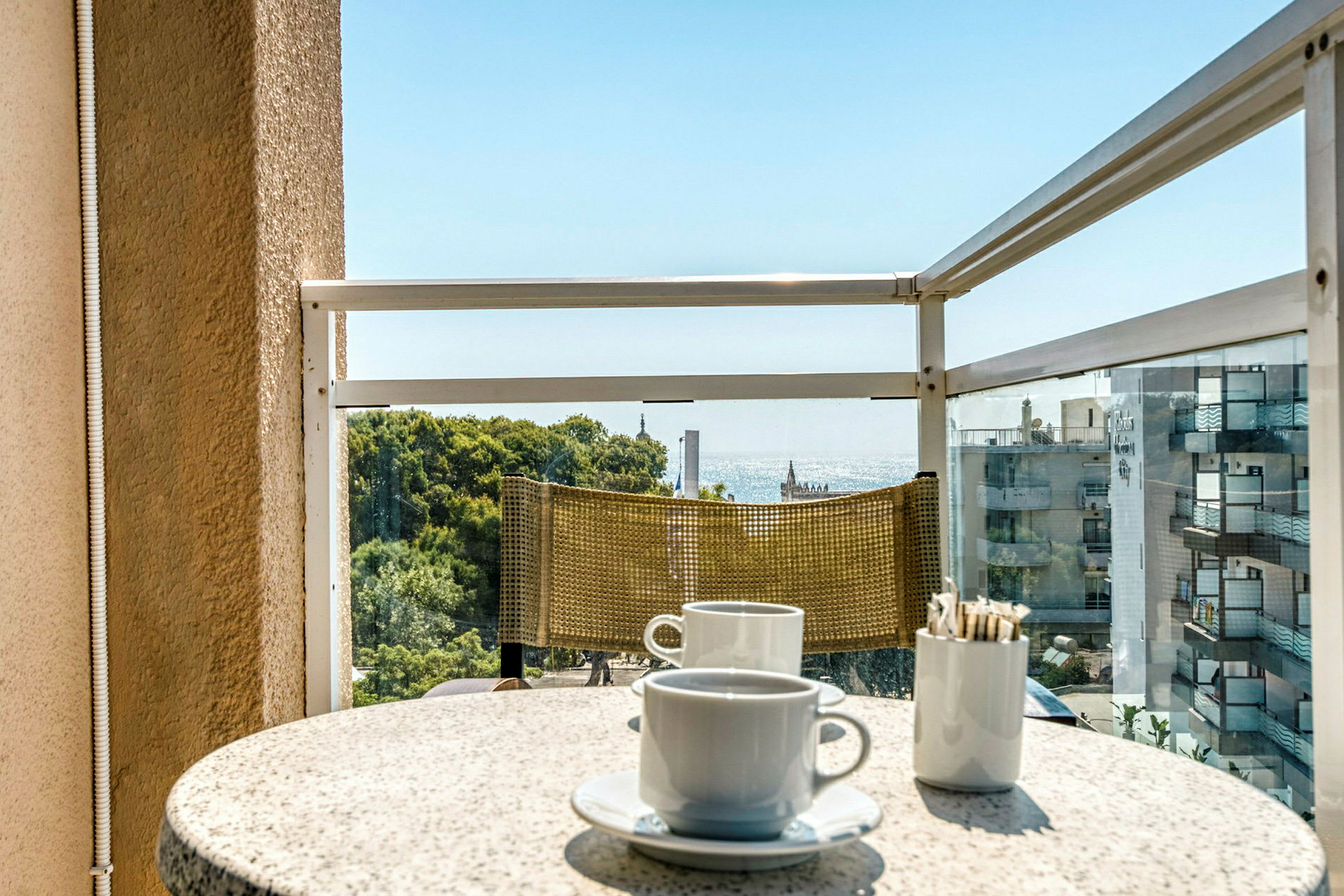 En balkong med utsikt över havet. På bordet finns två kaffekoppar och en behållare med sockersticks. (AI-genererad text)