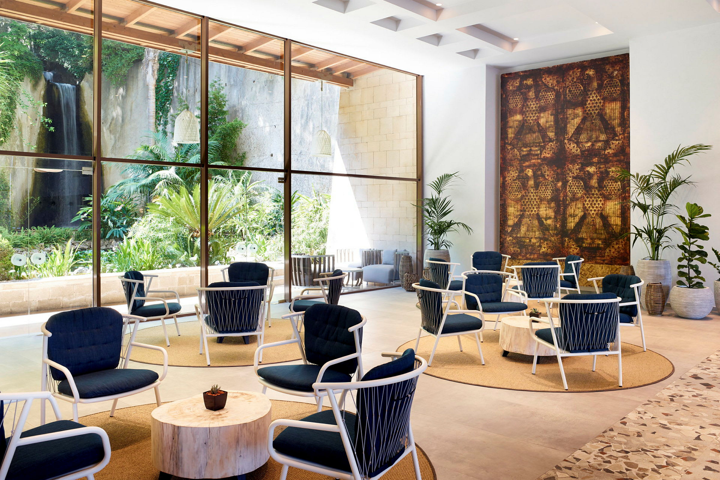 Hotelllobby med stolar och bord framför stora fönster med utsikt mot en trädgård och vattenfall. (AI-genererad text)