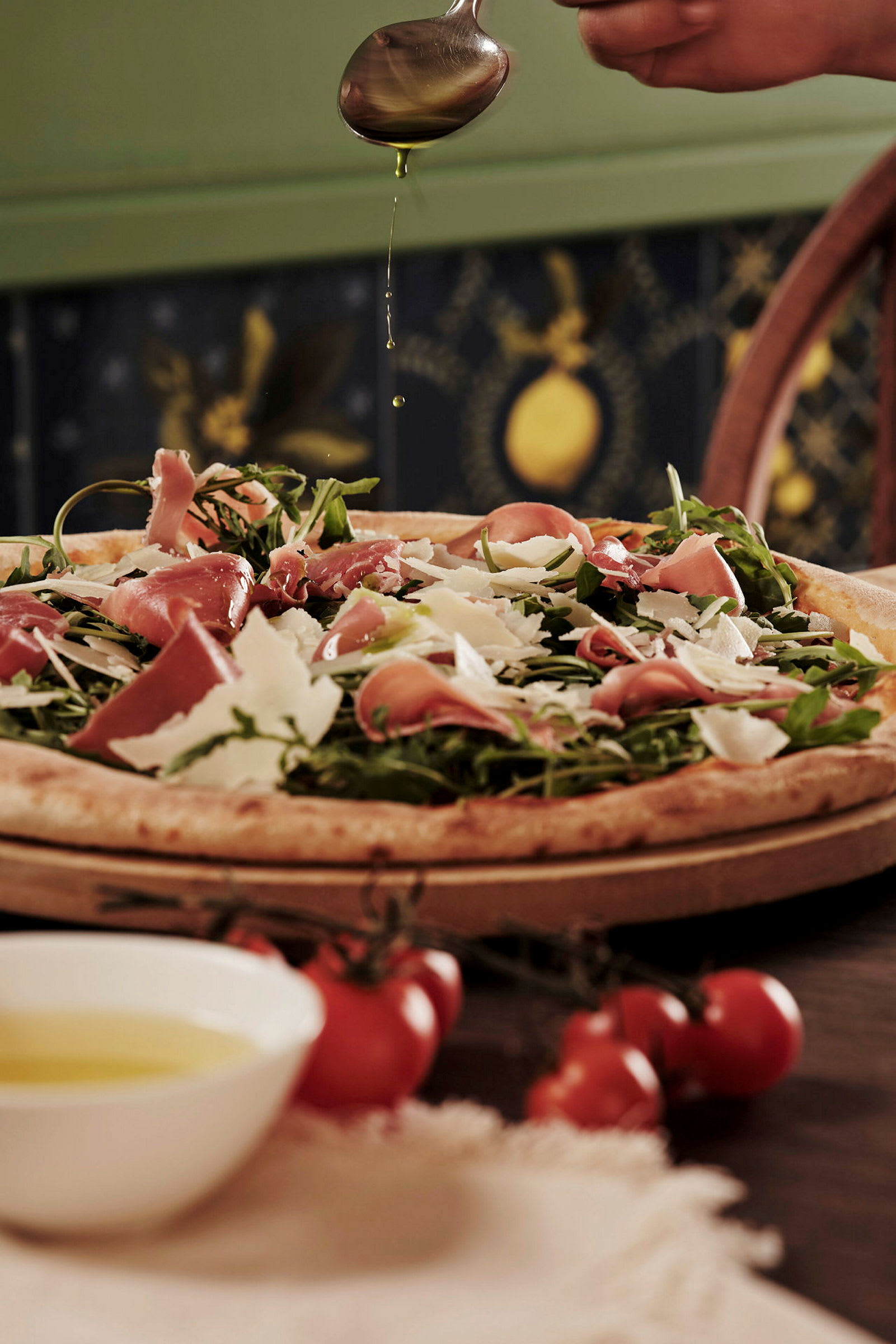 En pizza toppad med skinka, ruccola och parmesan, en hand droppar olivolja över den. (AI-genererad text)