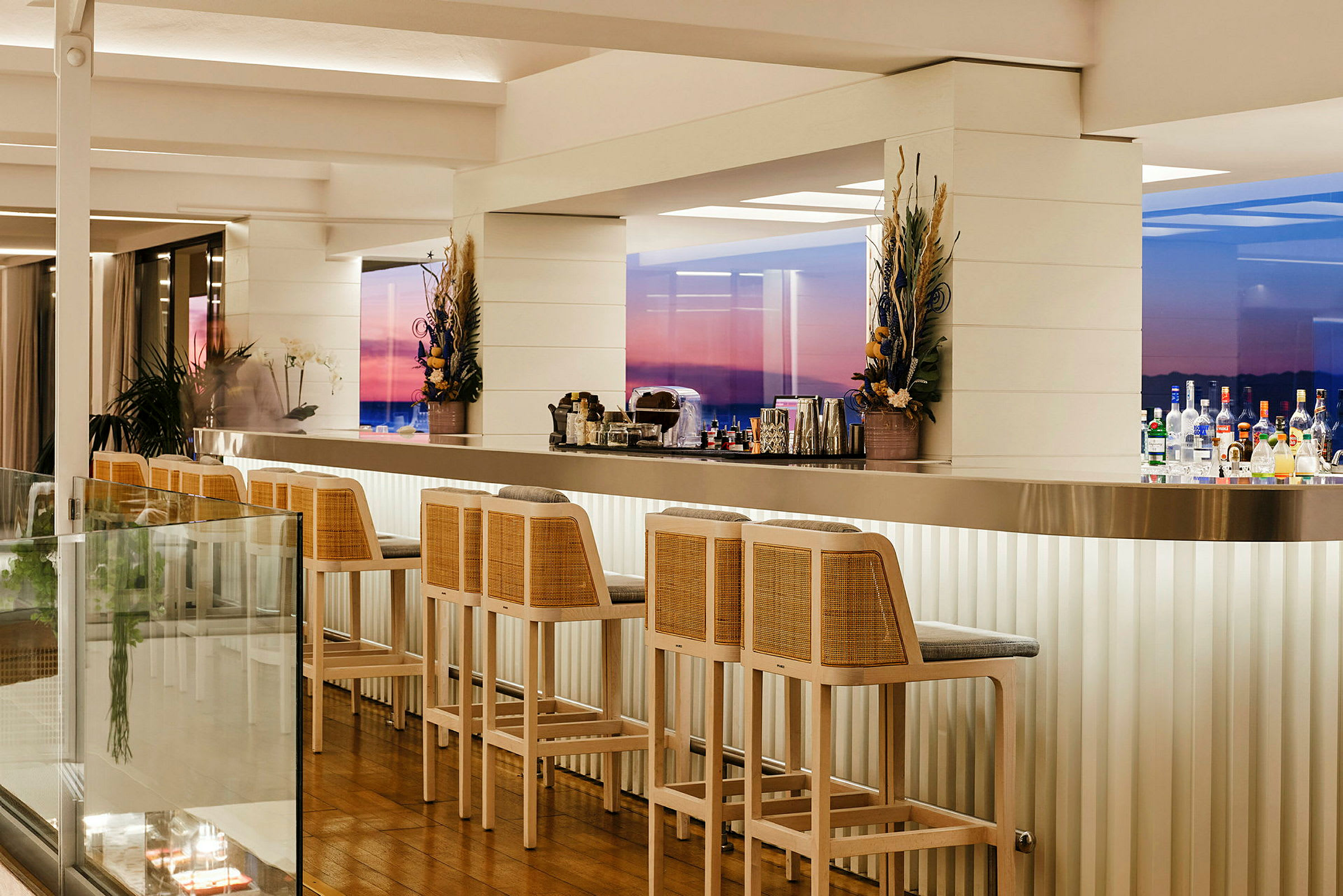 Bild av en stilfull bar med höga barstolar och en rad flaskor på diskbänken. Fönster i bakgrunden visar en färgstark solnedgång. (AI-genererad text)