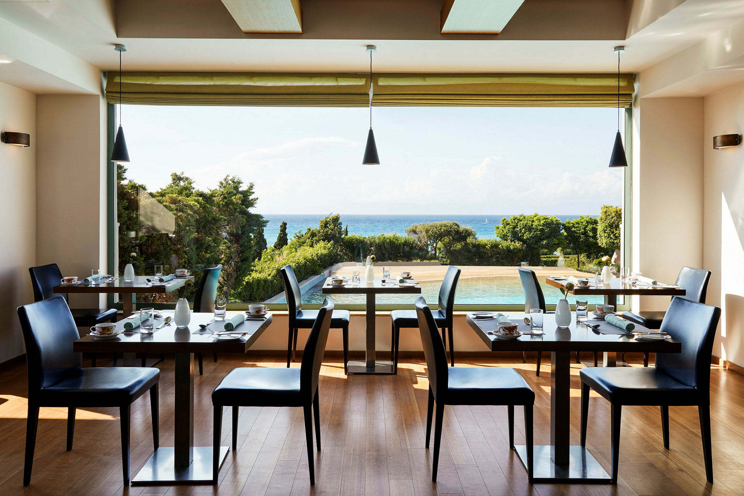 Ett ljust restaurangrum med uppdukade bord och stolar, med utsikt över en pool, gröna träd och havet i bakgrunden. (AI-genererad text)