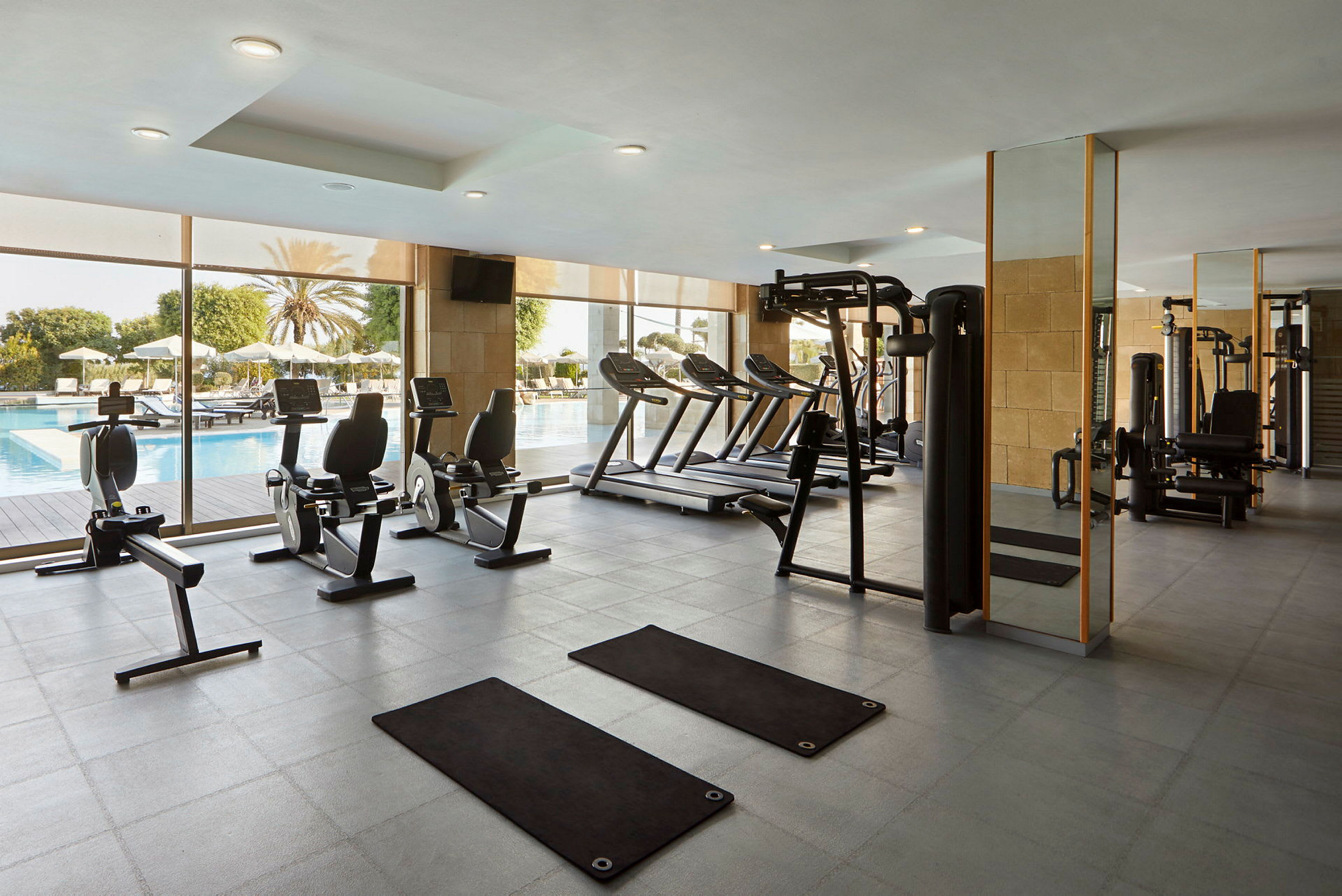 Ett gym med träningsutrustning och fönster mot en poolområde. (AI-genererad text)
