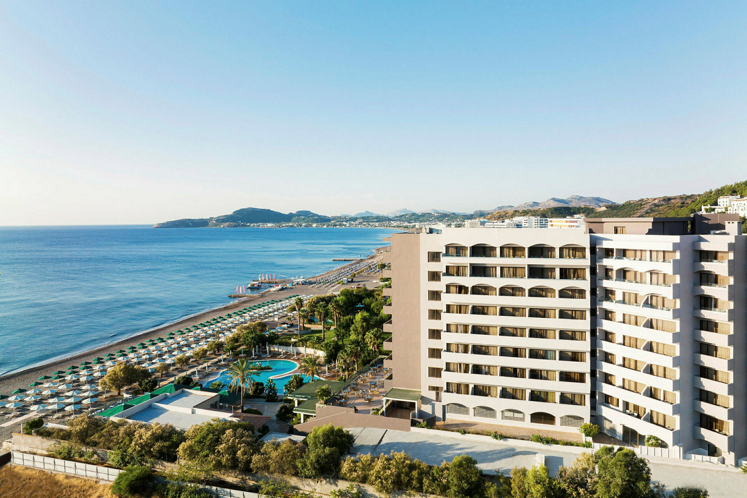 Vy över hotellbyggnad nära sandstrand och hav med solstolar och parasoller. (AI-genererad text)