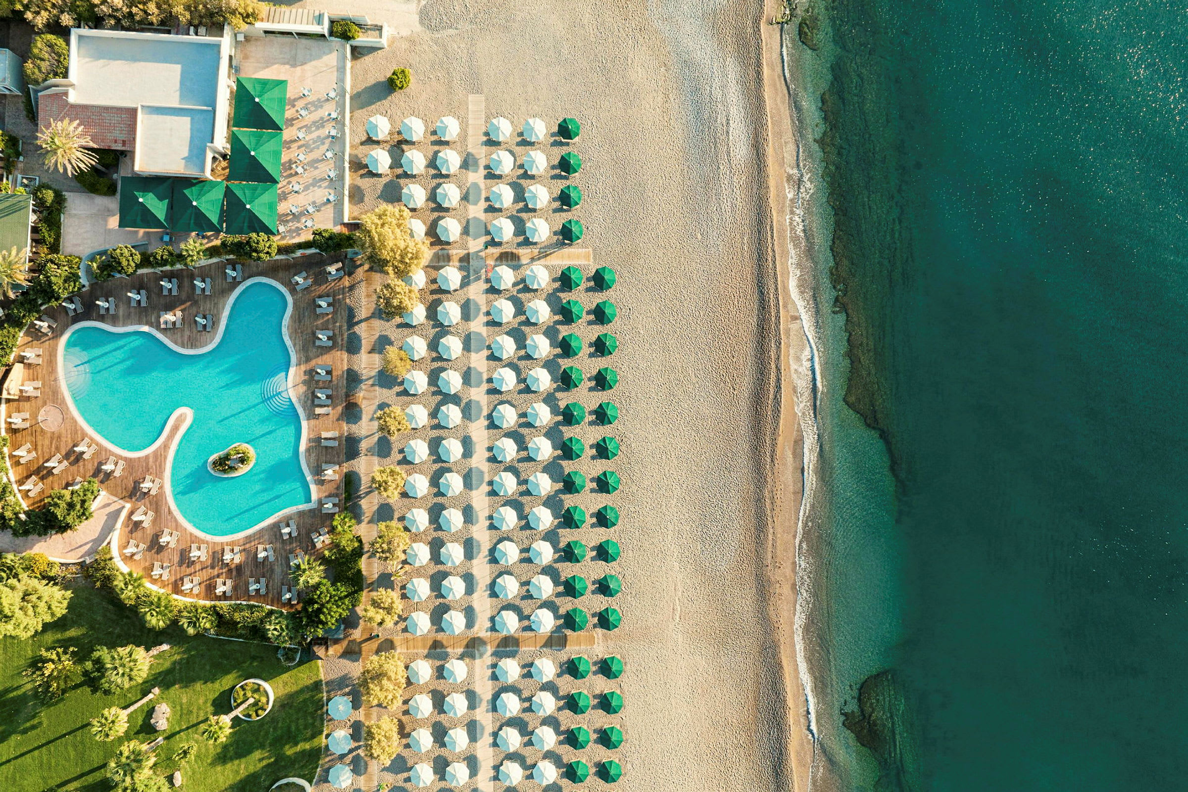 Flygfoto av en hotellpool med omgivande solstolar, bredvid en strand med parasoller och hav. (AI-genererad text)