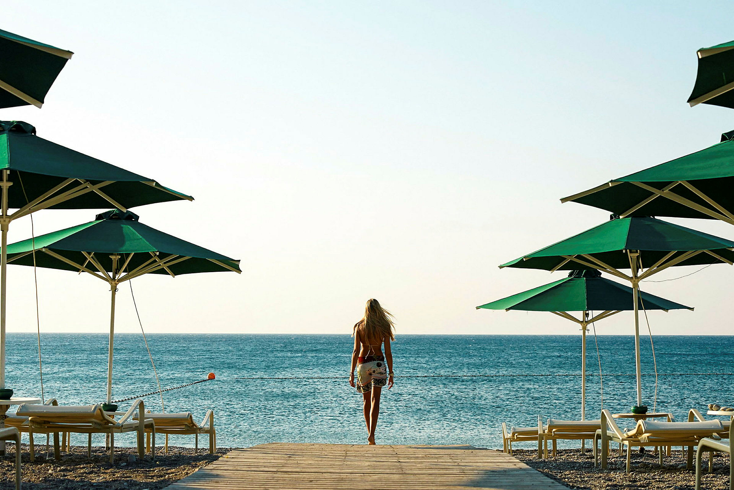 En person går mot havet mellan grönklädda parasoller och solstolar på stranden. (AI-genererad text)