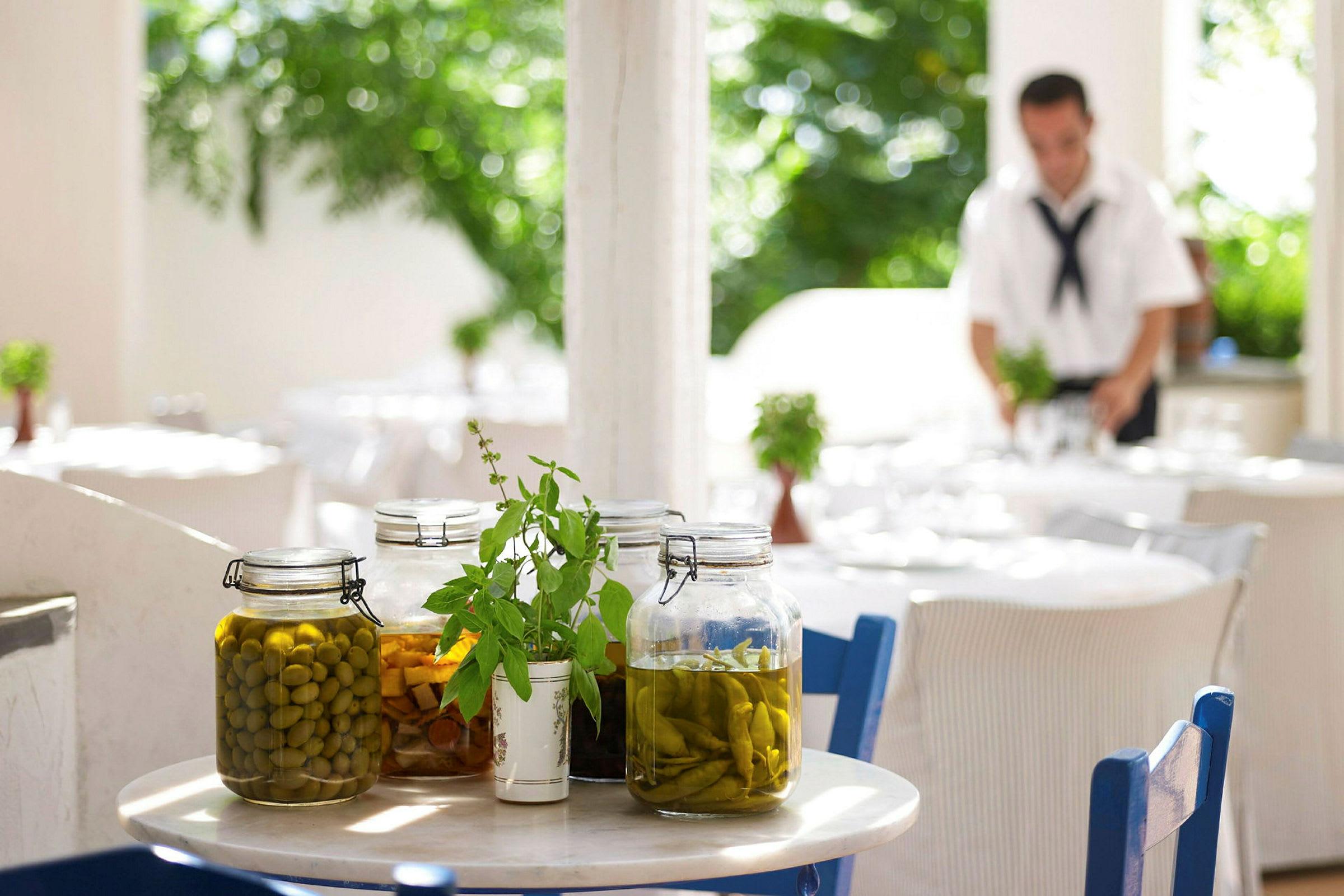 Ett bord med glasburkar fyllda med inlagda grönsaker och örtdekorationer. Bakgrunden visar en utomhusrestaurang. (AI-genererad text)