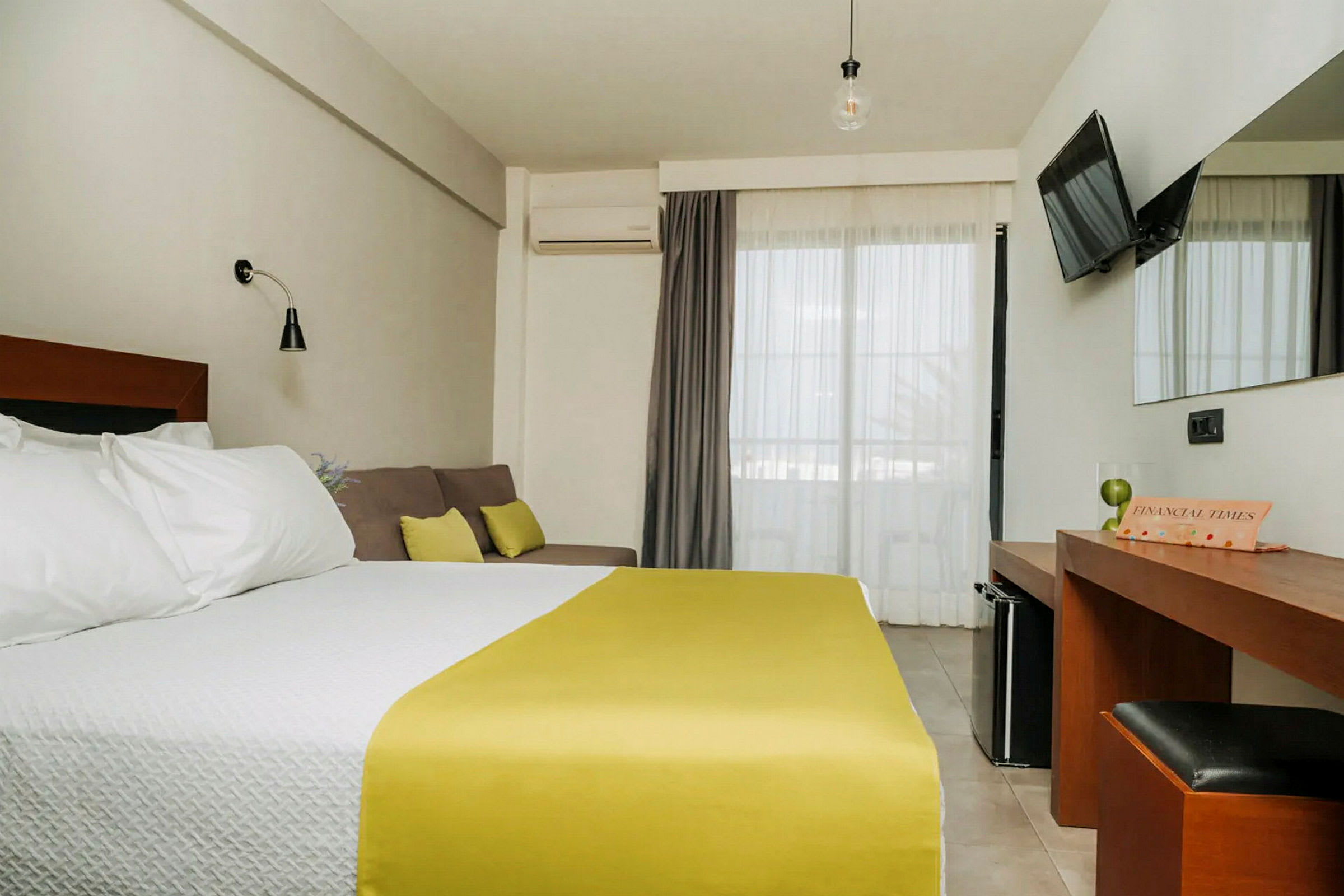 Ett hotellrum med en dubbelsäng med vita sängkläder och gult överkast, en soffa med kuddar, ett skrivbord med spegel, och en TV på väggen. Balkongdörrar med ljusa gardiner. (AI-genererad text)