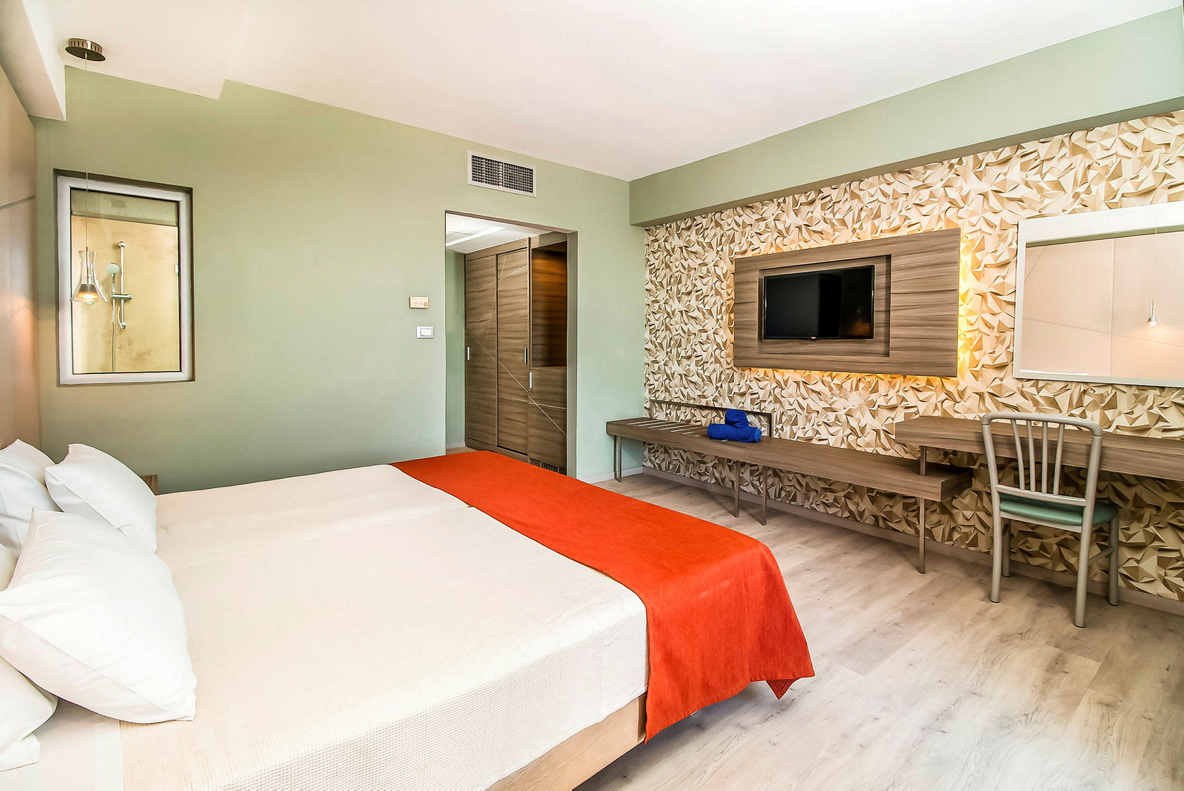 Hotellrum med en dubbelsäng täckt med vit och orange sängkläder, en vägg med geometrisk dekor, samt en tv och skrivbord. (AI-genererad text)