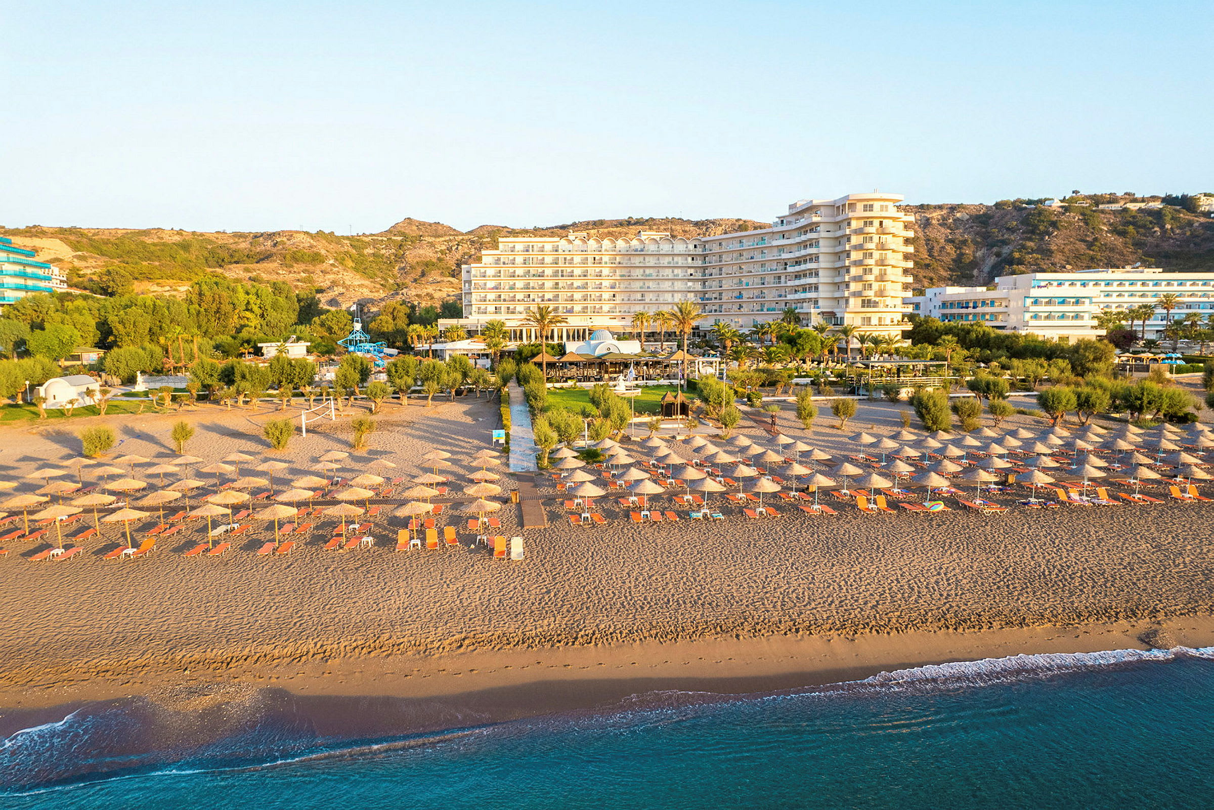 Strand med solparasoller och solstolar, hotell och grönska i bakgrunden. (AI-genererad text)