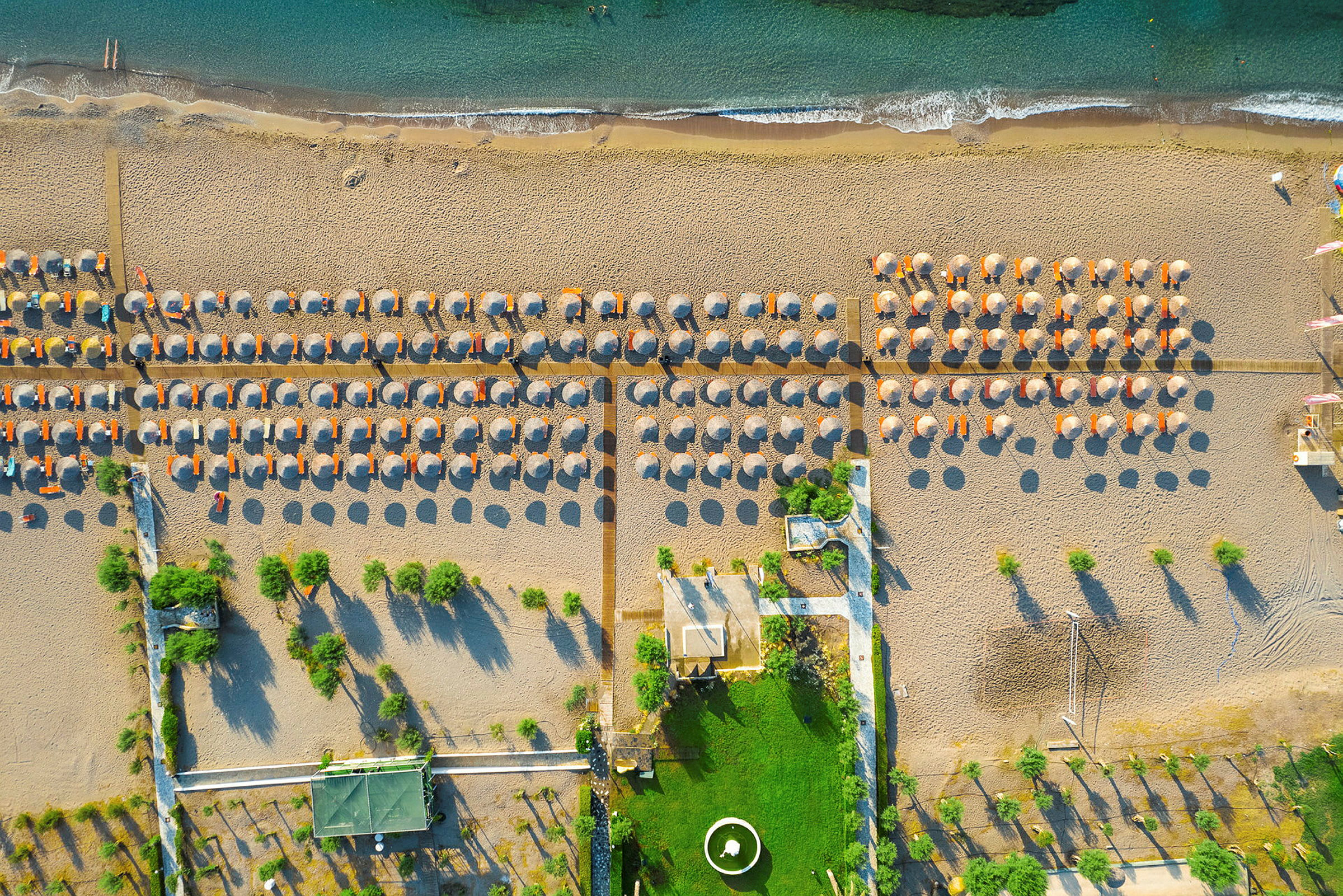 Flygfoto: Sandstrand med raden av solstolar och parasoller bredvid grönyta och hav. (AI-genererad text)