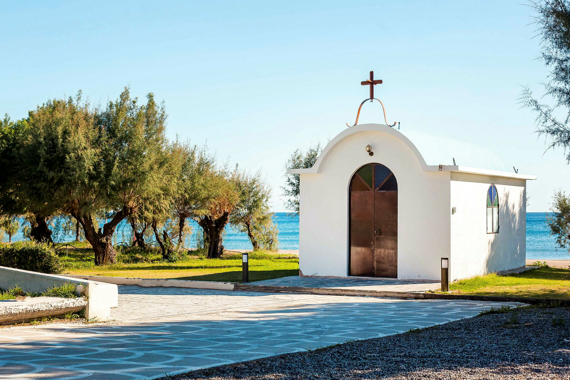 En vit kyrka med kopparkors och glasfönster, omgiven av gröna träd vid havet. (AI-genererad text)