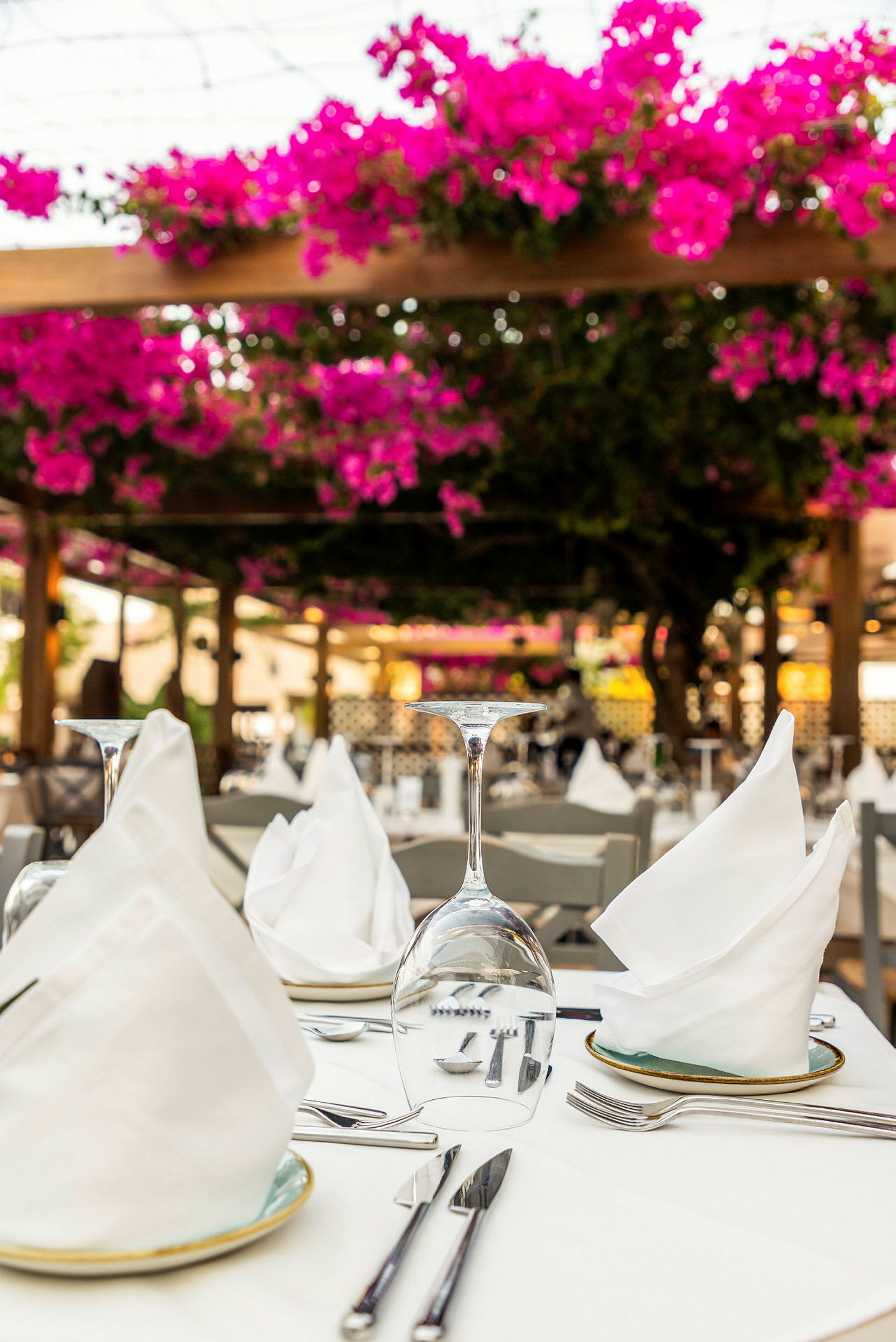 Ett dukat bord i en utomhusrestaurang med vita servetter och vinglas, omgärdat av rosa blommor. (AI-genererad text)