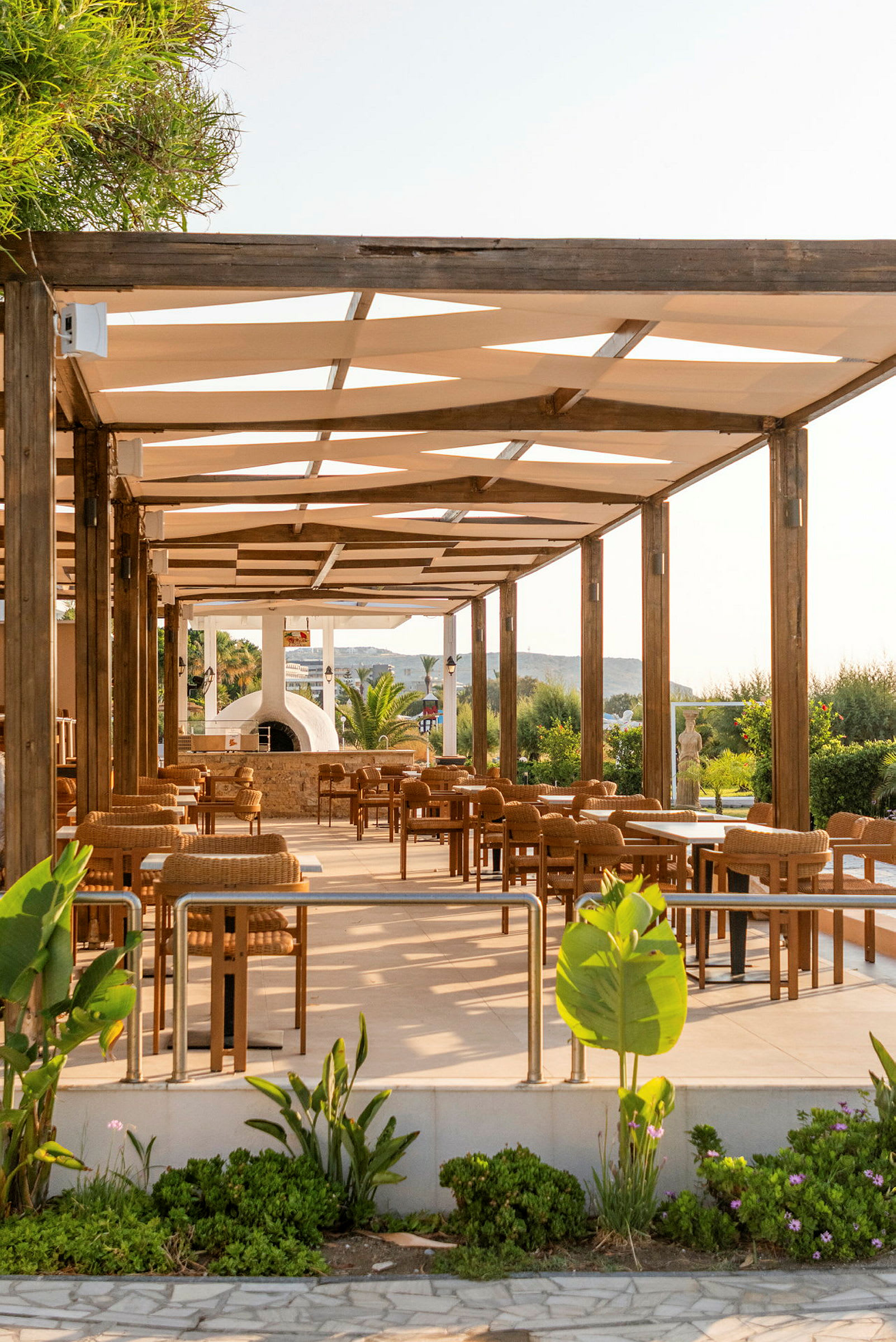 En utomhusrestaurang med trästolar och bord under en pergola, omgiven av grönska. (AI-genererad text)