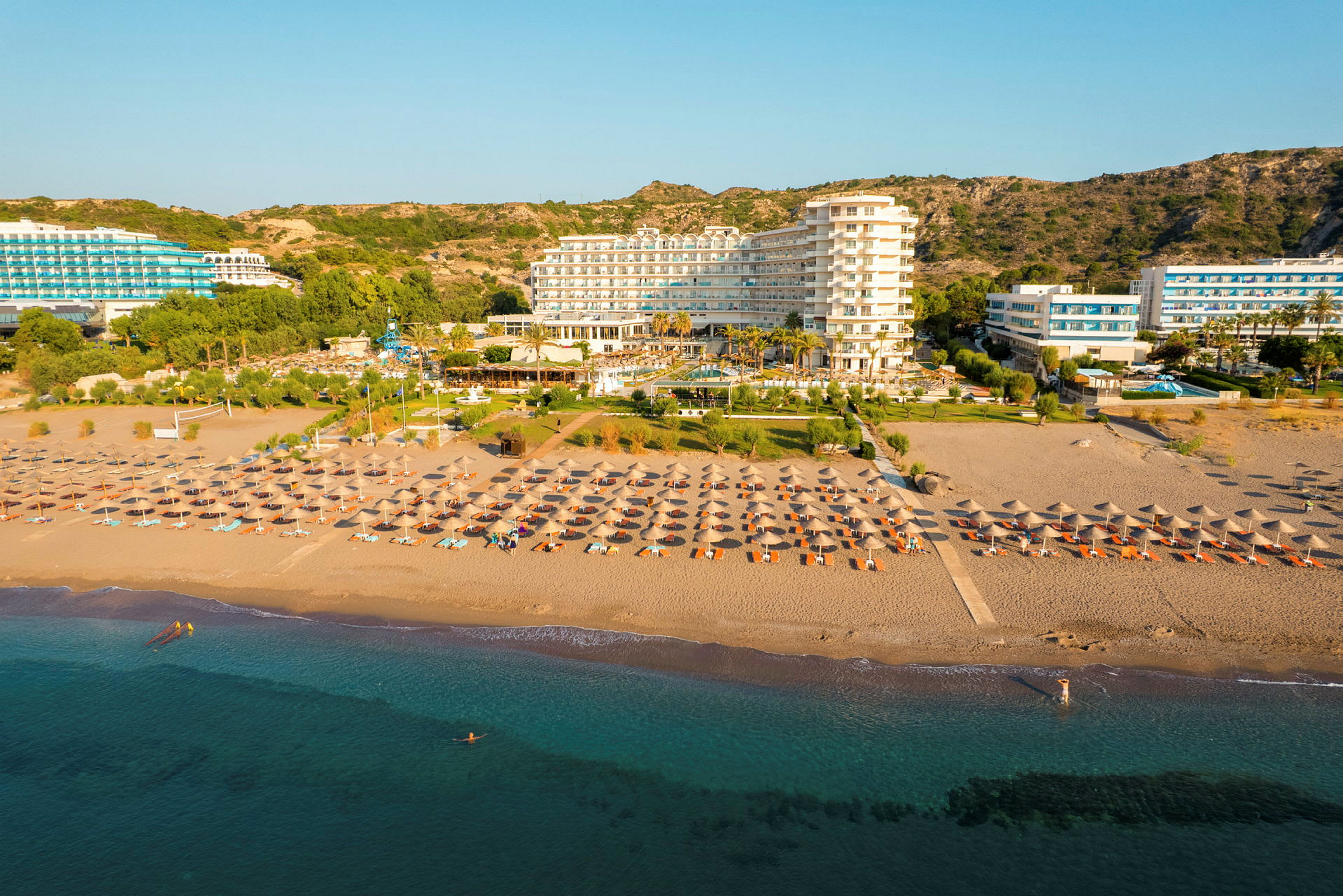 Ett hotell vid stranden med solstolar och parasoller framför och en klar blå himmel i bakgrunden. (AI-genererad text)
