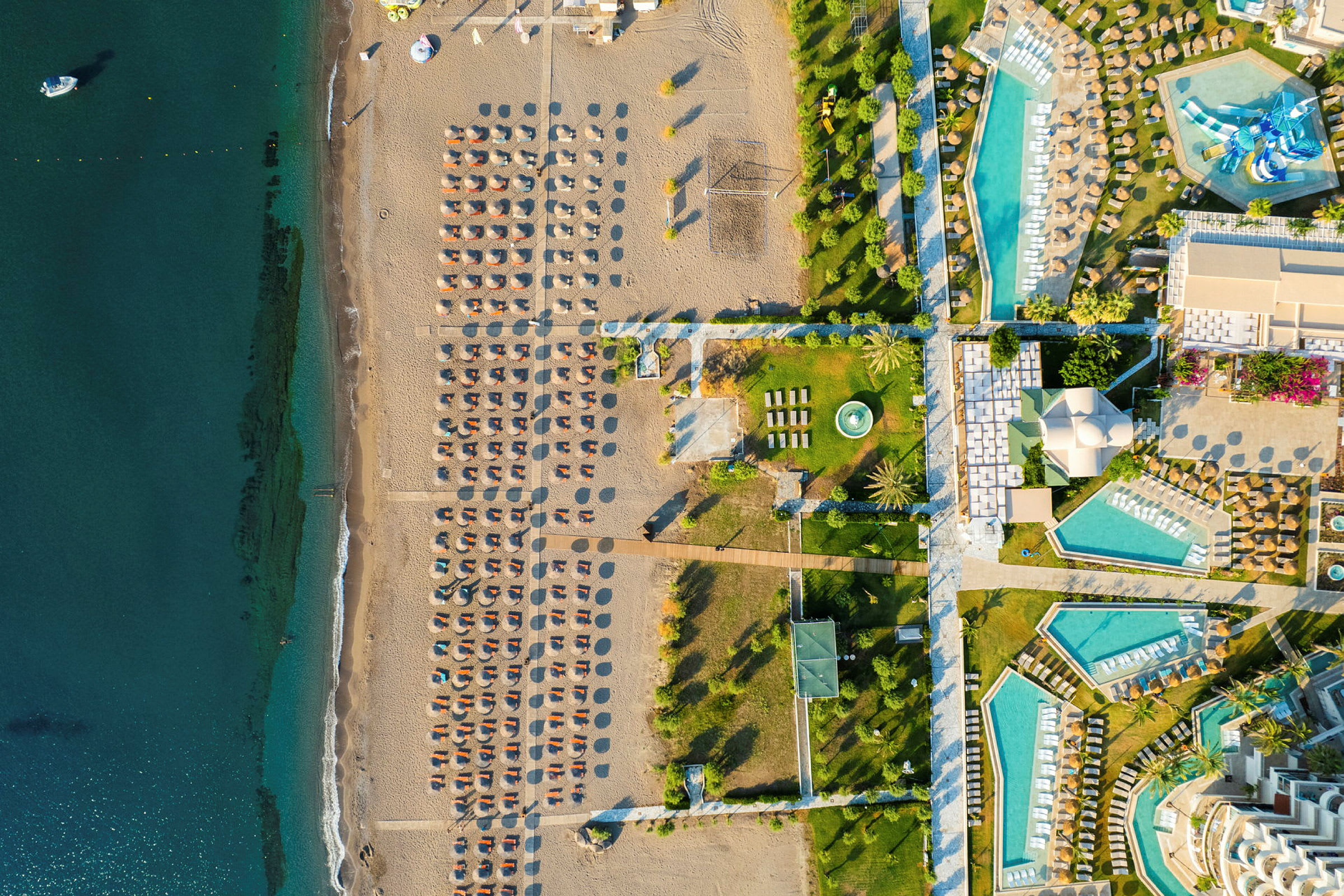 Flygbild över en strand med solstolar och parasoller bredvid ett hotellområde med pooler och grönområden. (AI-genererad text)