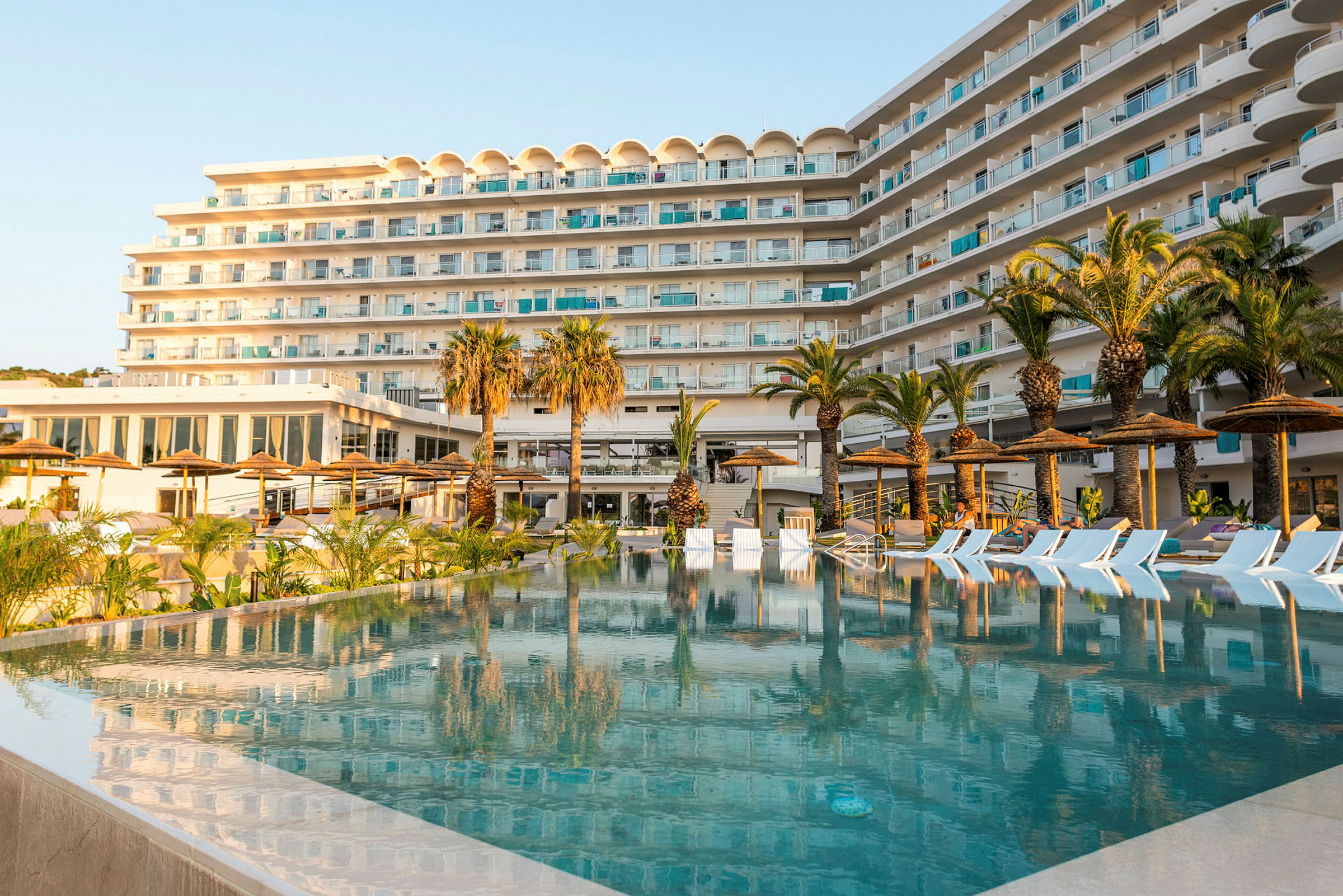 Ett hotell med flera våningar framför en pool omgiven av palmer och solstolar. (AI-genererad text)