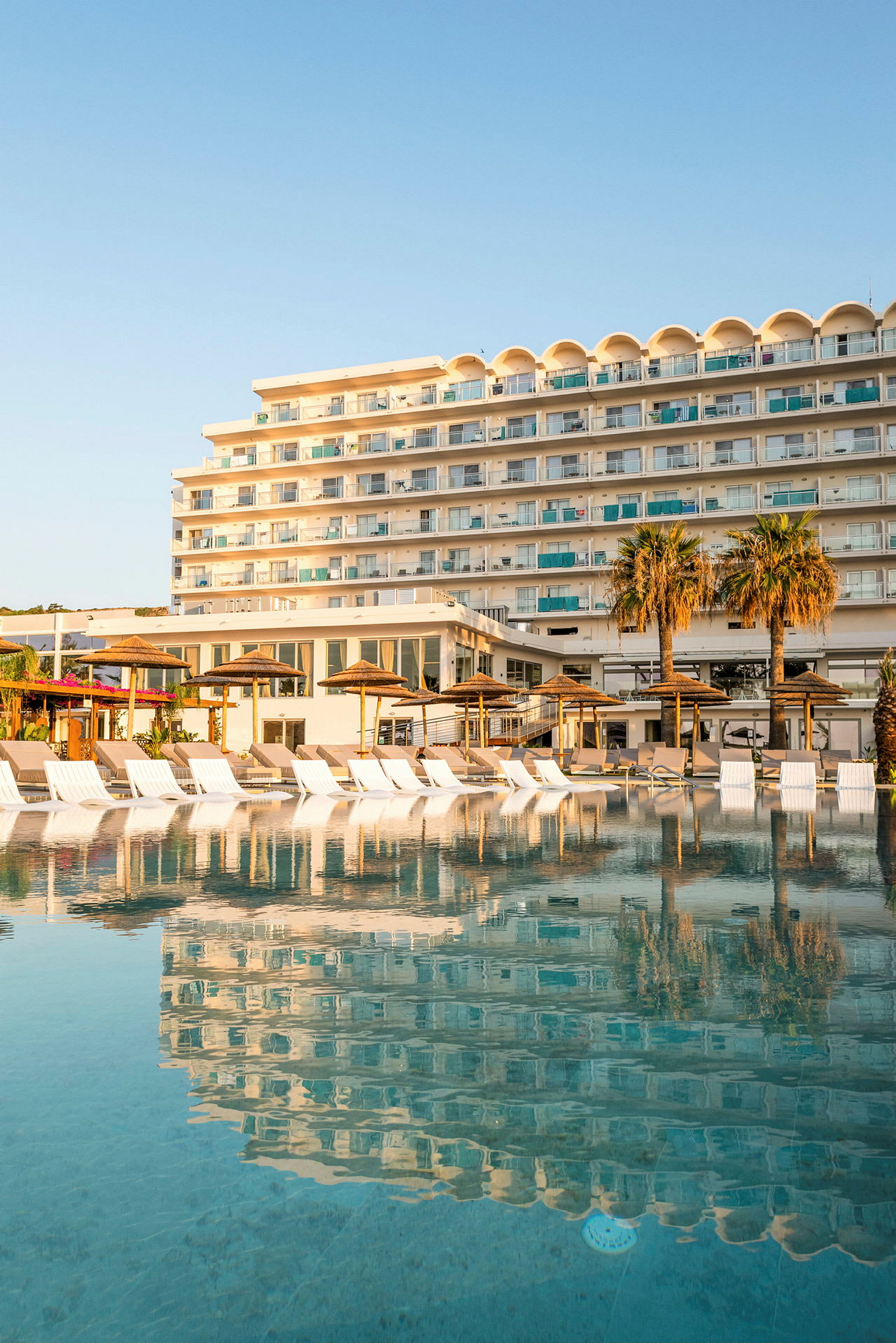 Hotellbyggnad vid en stor pool med solstolar och parasoller i förgrunden. (AI-genererad text)