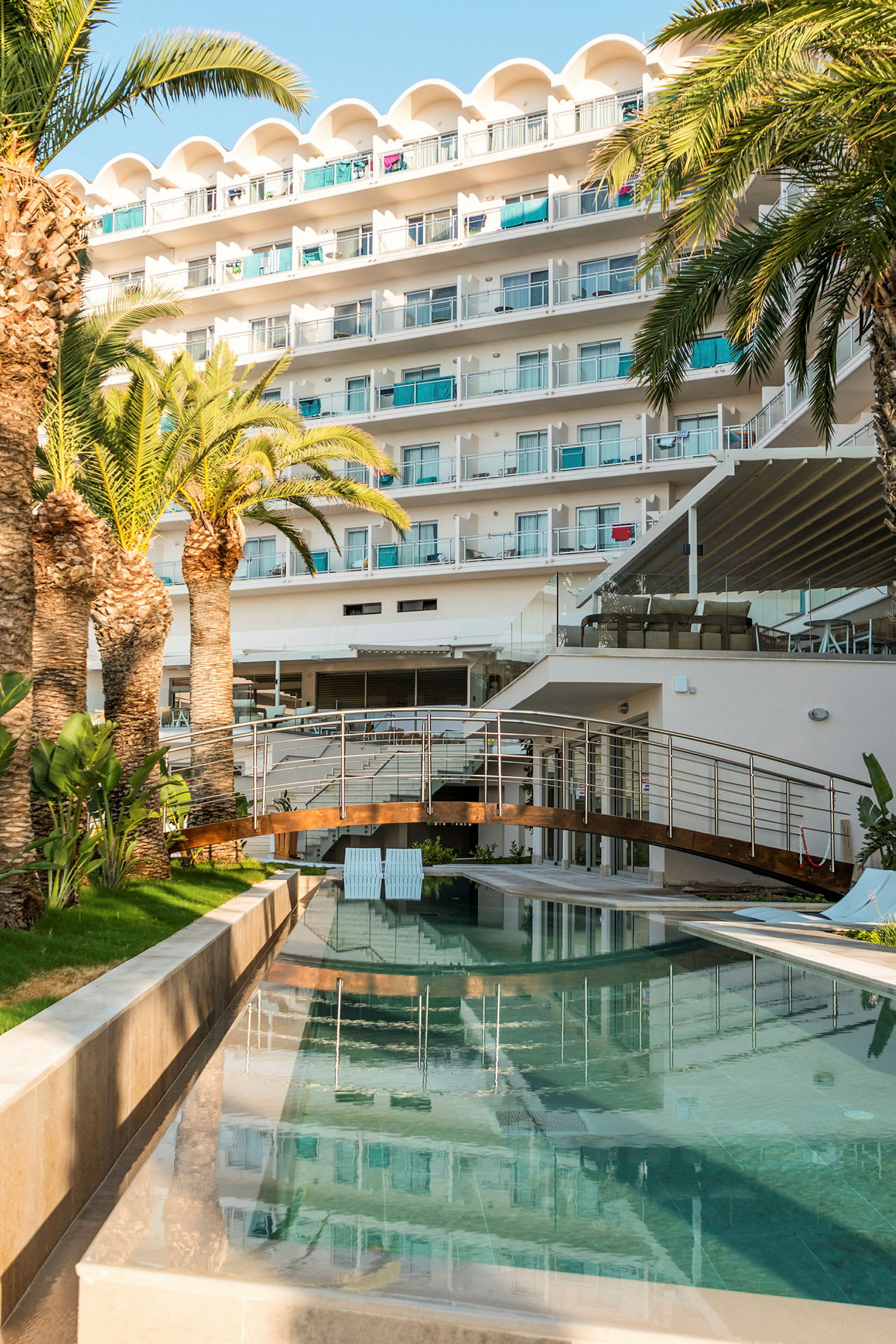 Ett hotellkomplex med flera våningar omgivet av palmer och träd. I förgrunden syns en pool och en gångbro. (AI-genererad text)