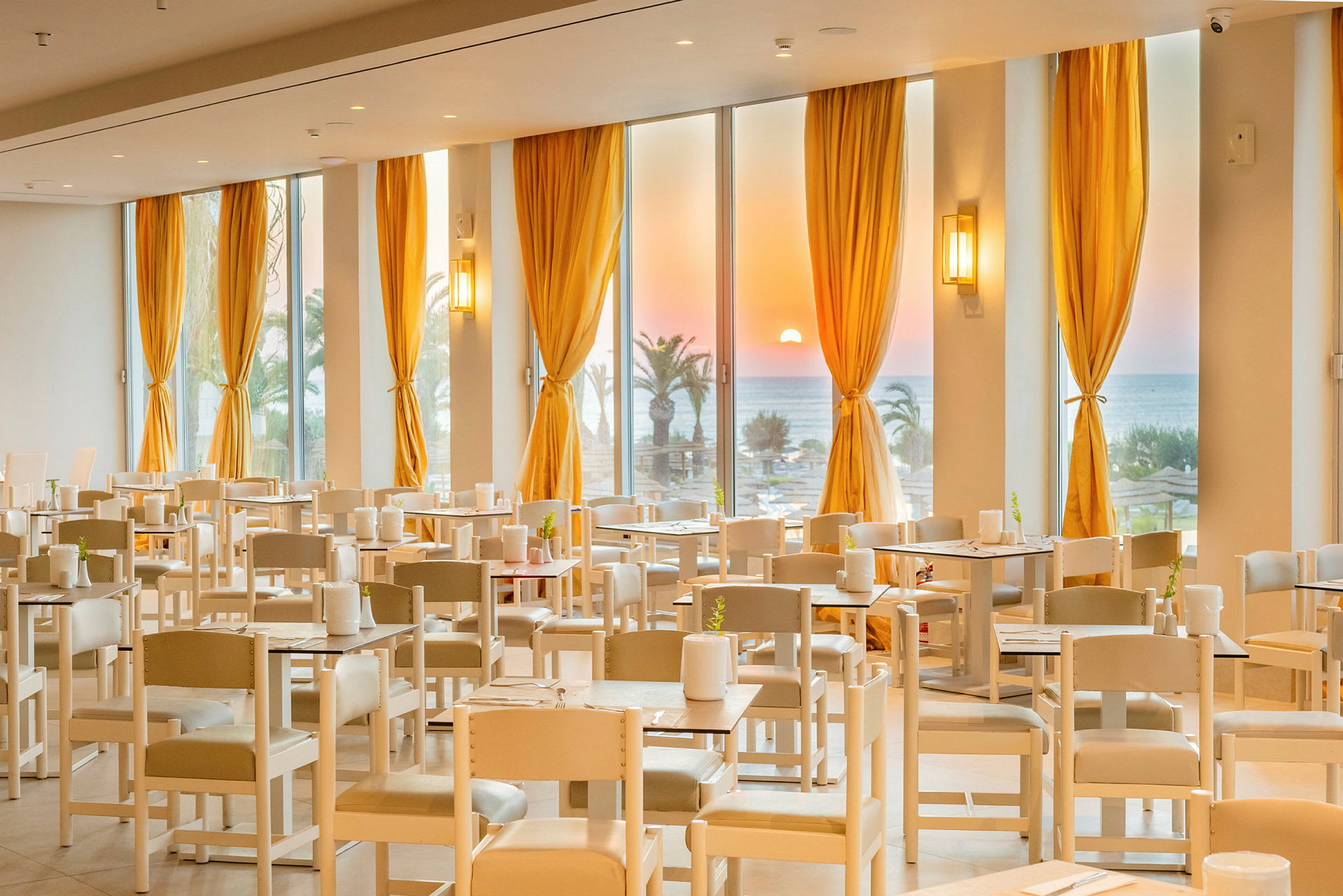 En restaurang med bordsuppsättningar och vita stolar. Stora fönster med gula gardiner visar en solnedgång över havet och palmer. (AI-genererad text)