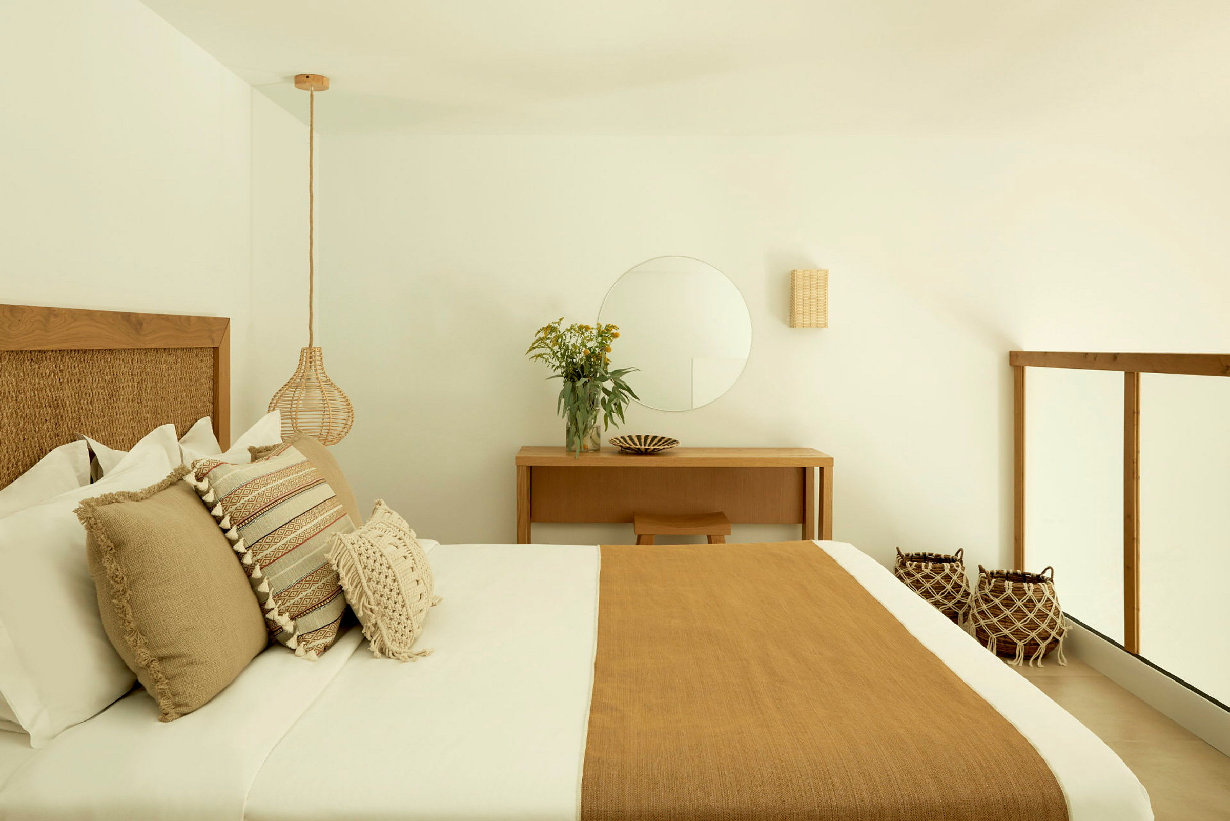 Ett elegant sovrum med en dubbelsäng, dekorerad med bruna, beige och vita kuddar samt en matchande sängsprid. Väggarna är vita med minimalistisk inredning och en spegel ovanför ett skrivbord. (AI-genererad text)