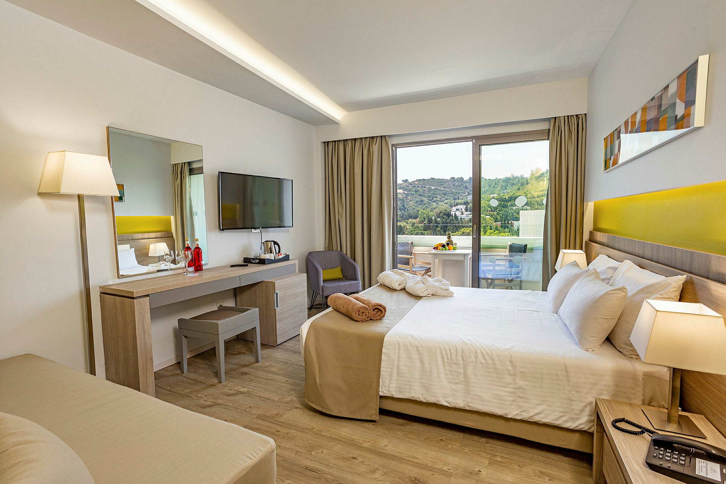 Ett hotellrum med säng, skrivbord, TV, och en balkong som visar en grön utsikt. (AI-genererad text)