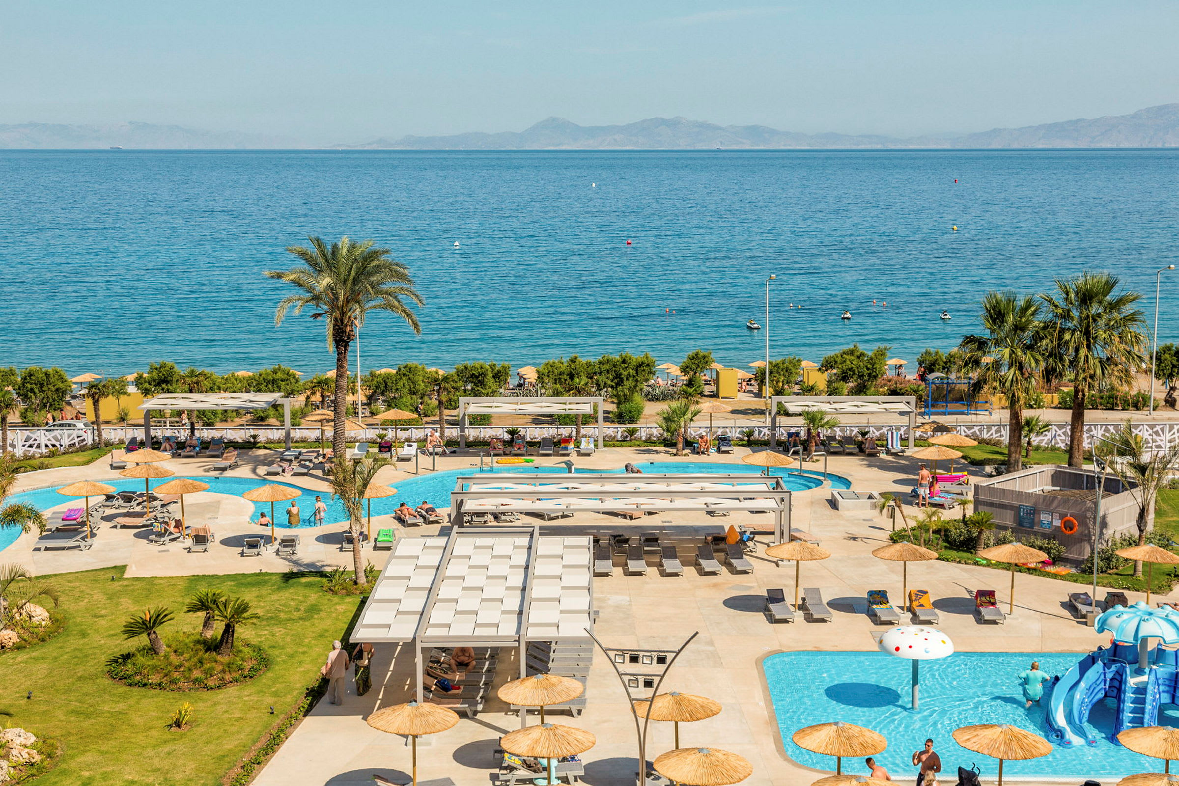 En vy över hotellområde med pooler och solstolar, havet syns i bakgrunden. (AI-genererad text)