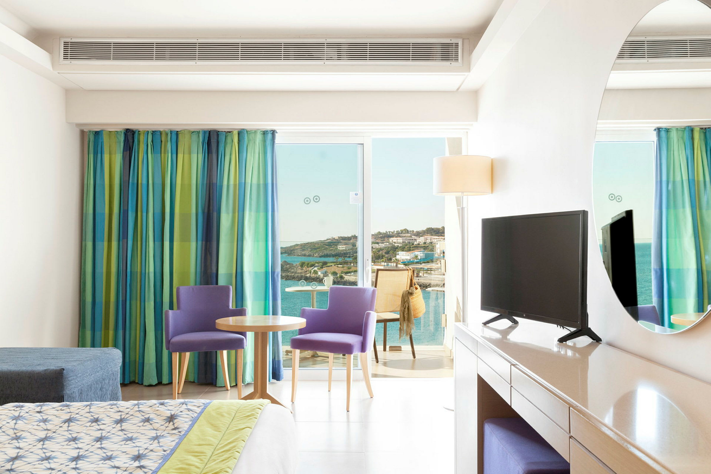 Ett hotellrum med säng, TV på byrå och sittgrupp framför en glasdörr som leder till balkong med utsikt över havet. (AI-genererad text)