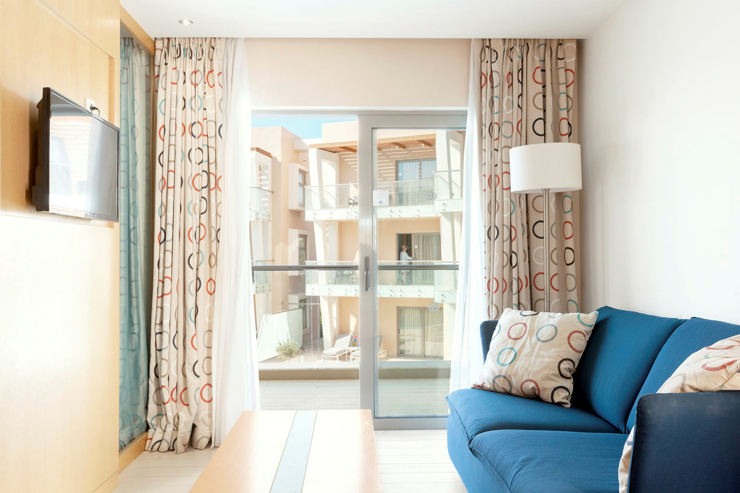 Ett ljust hotellrum med blå soffa, cirkelformade gardiner och utgång till en balkong. (AI-genererad text)
