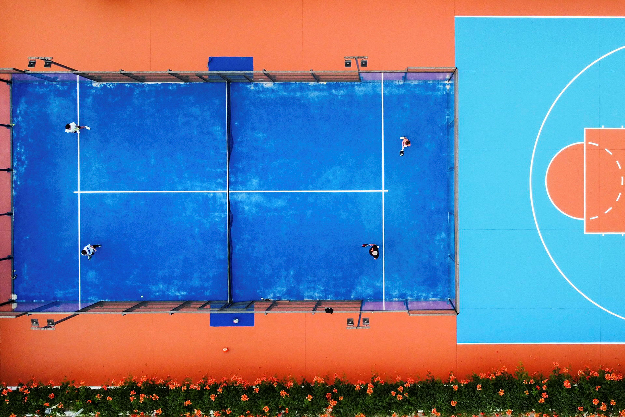 Luftvy av en blå padelbana med människor som spelar, bredvid en orange och blå basketplan. (AI-genererad text)