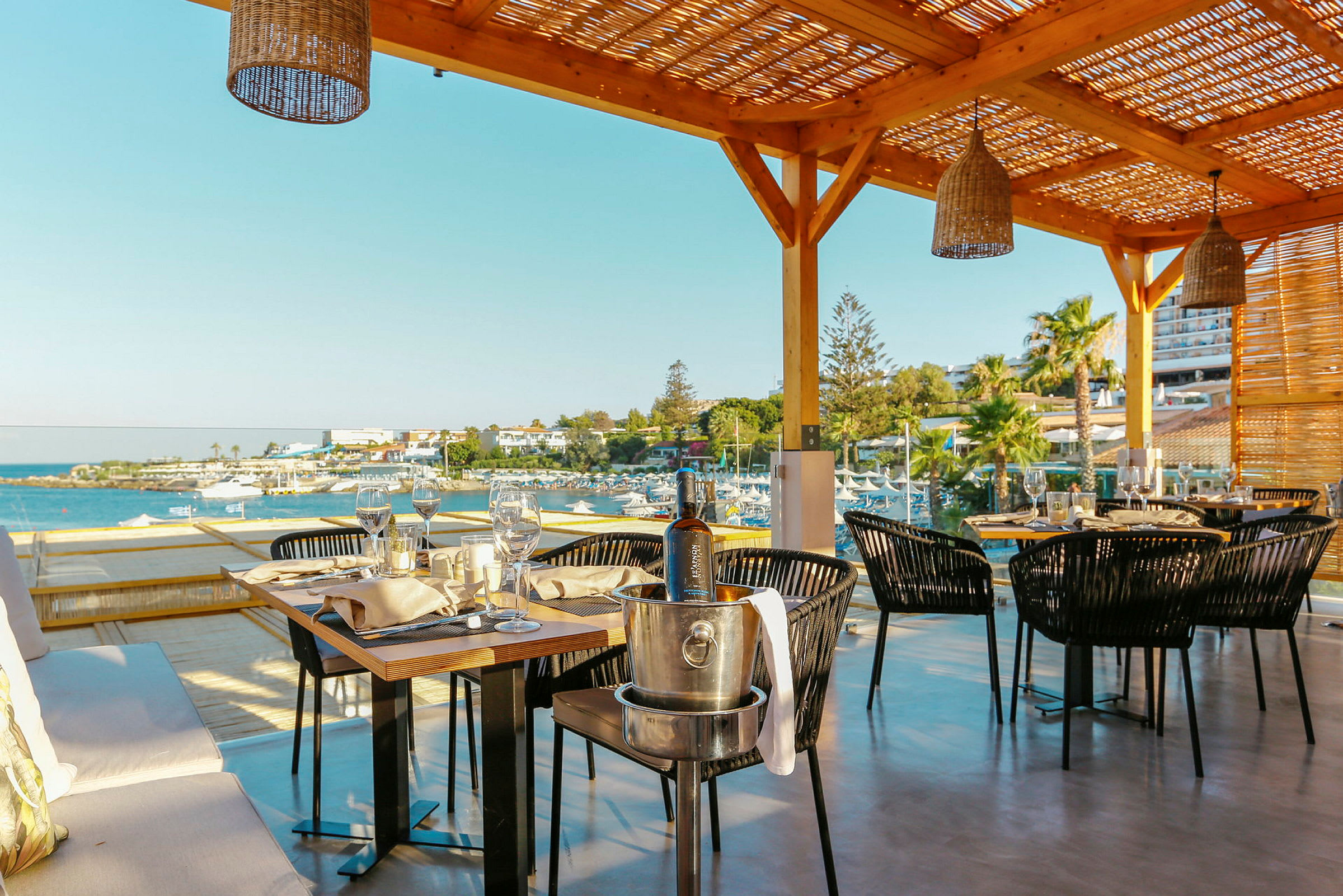 Uteservering med bord och stolar under en pergola, med utsikt över hav och strand. (AI-genererad text)