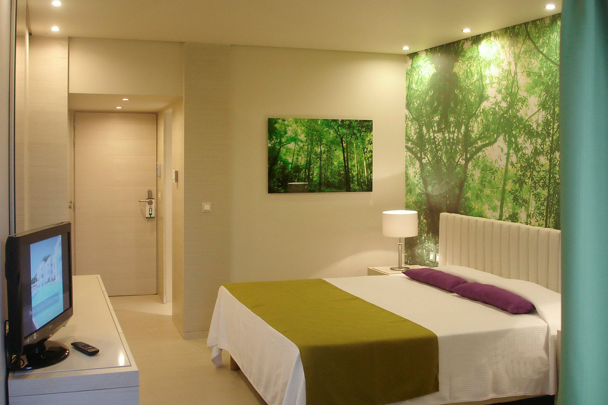 Ett hotellrum med en säng, grön och vit inredning, en TV och en väggdekoration föreställande en skog. (AI-genererad text)