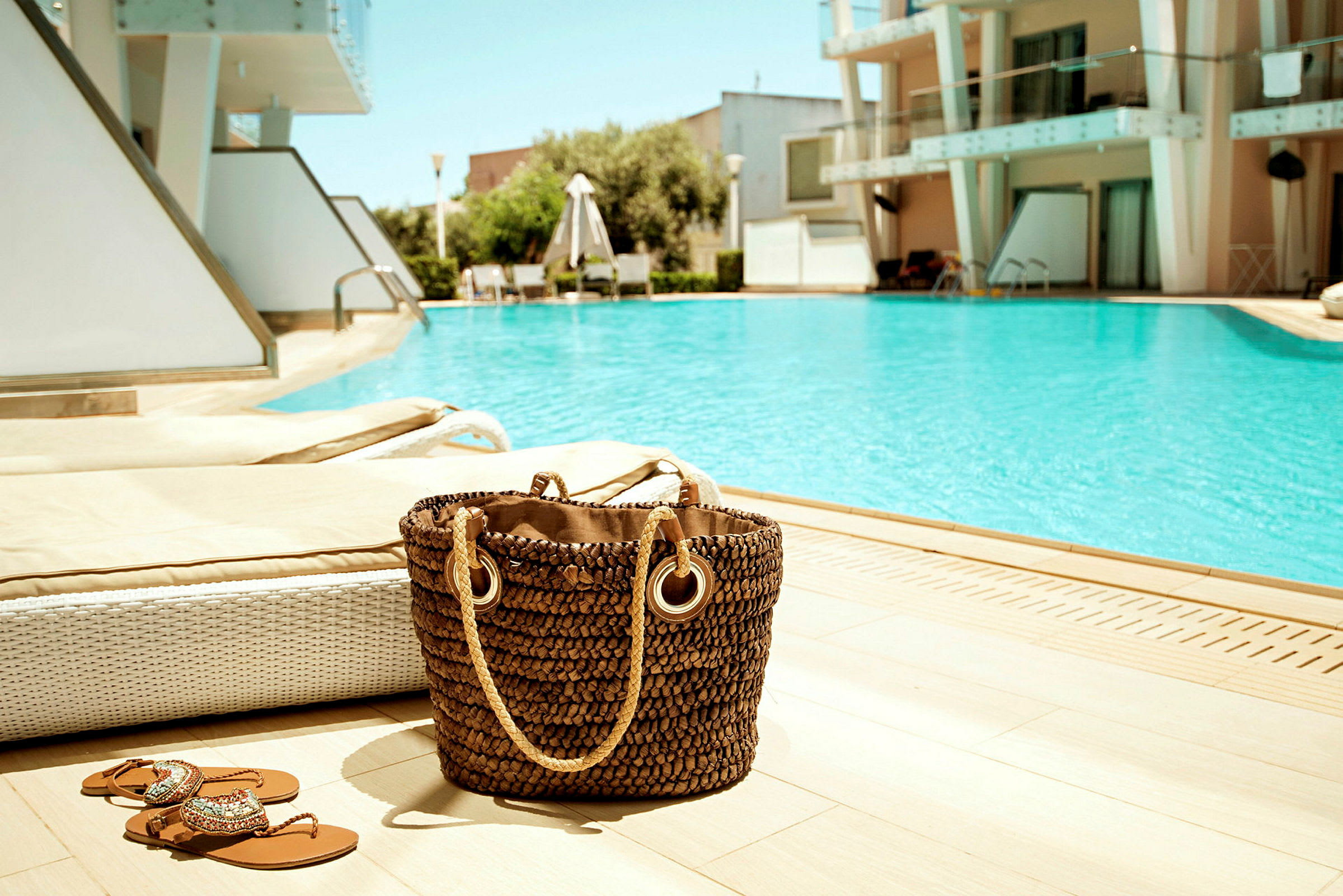 Strandväska och sandaler vid en pool, omgiven av solstolar och en modern byggnad i bakgrunden. (AI-genererad text)