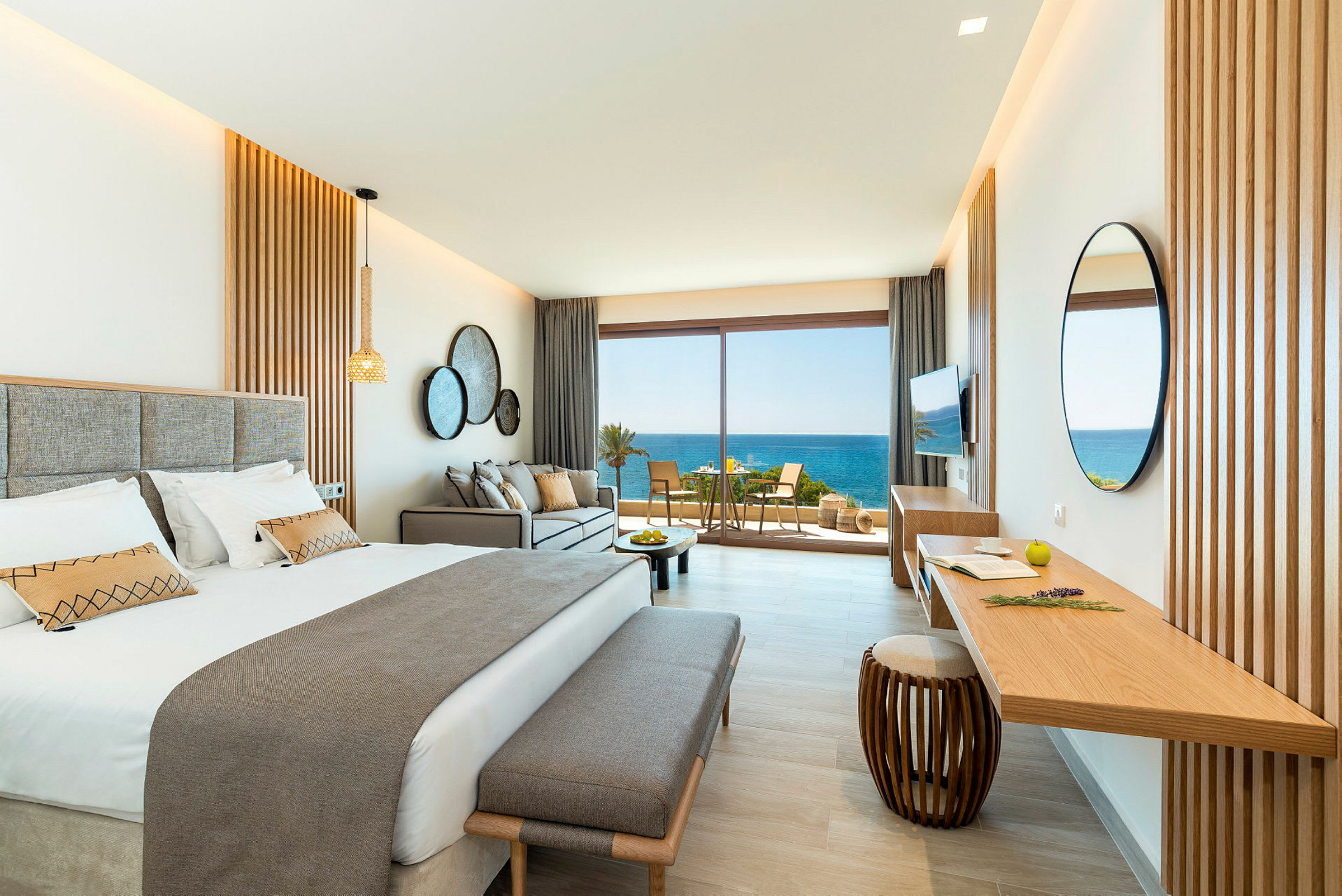 Ett stilrent hotellrum med en dubbelsäng, soffa och ett skrivbord. Stora glasdörrar öppnar upp till en balkong med utsikt över havet. (AI-genererad text)