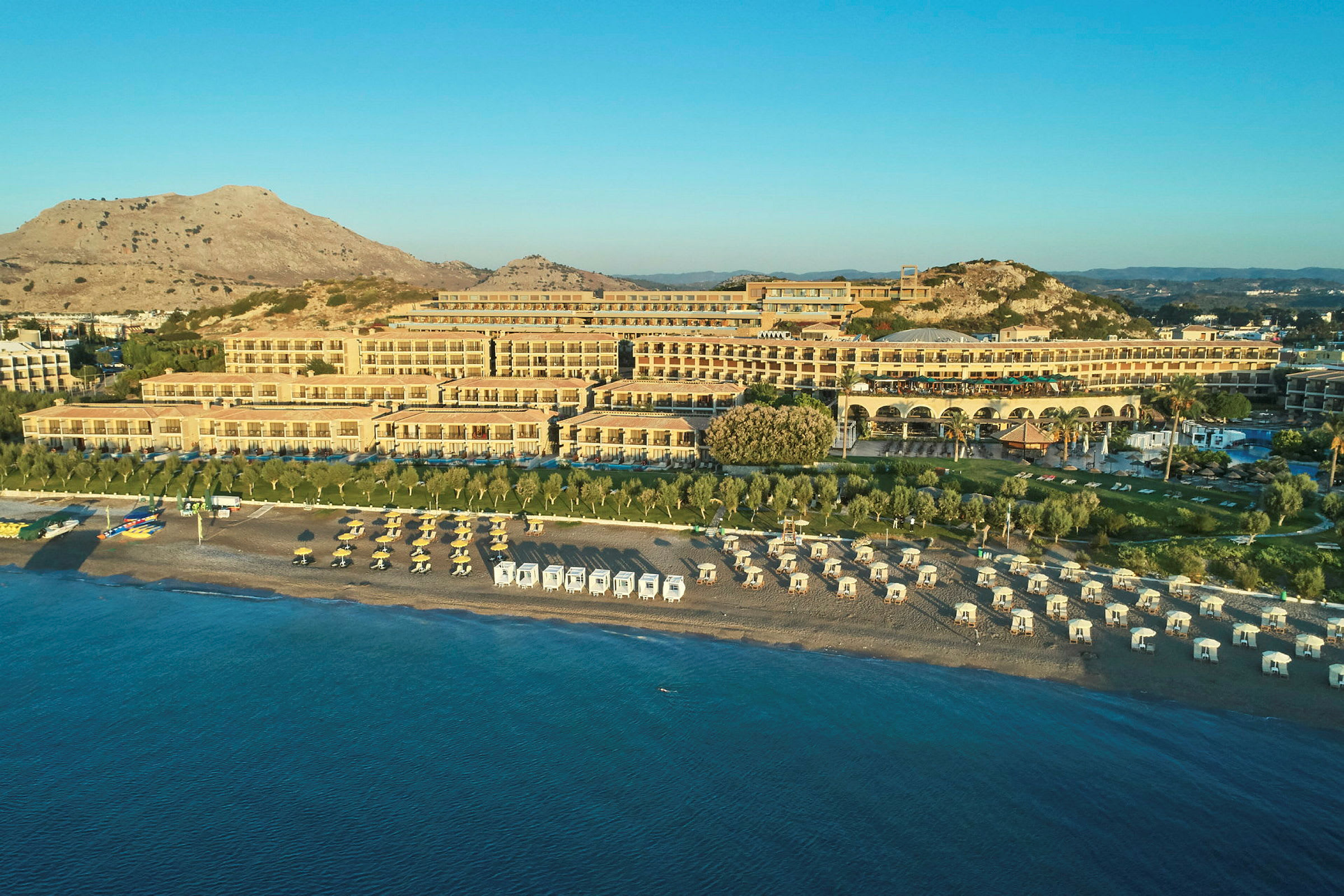 Vid en solig kust ligger ett stort hotell med flera terrasser och rader av solstolar och parasoller på stranden. (AI-genererad text)