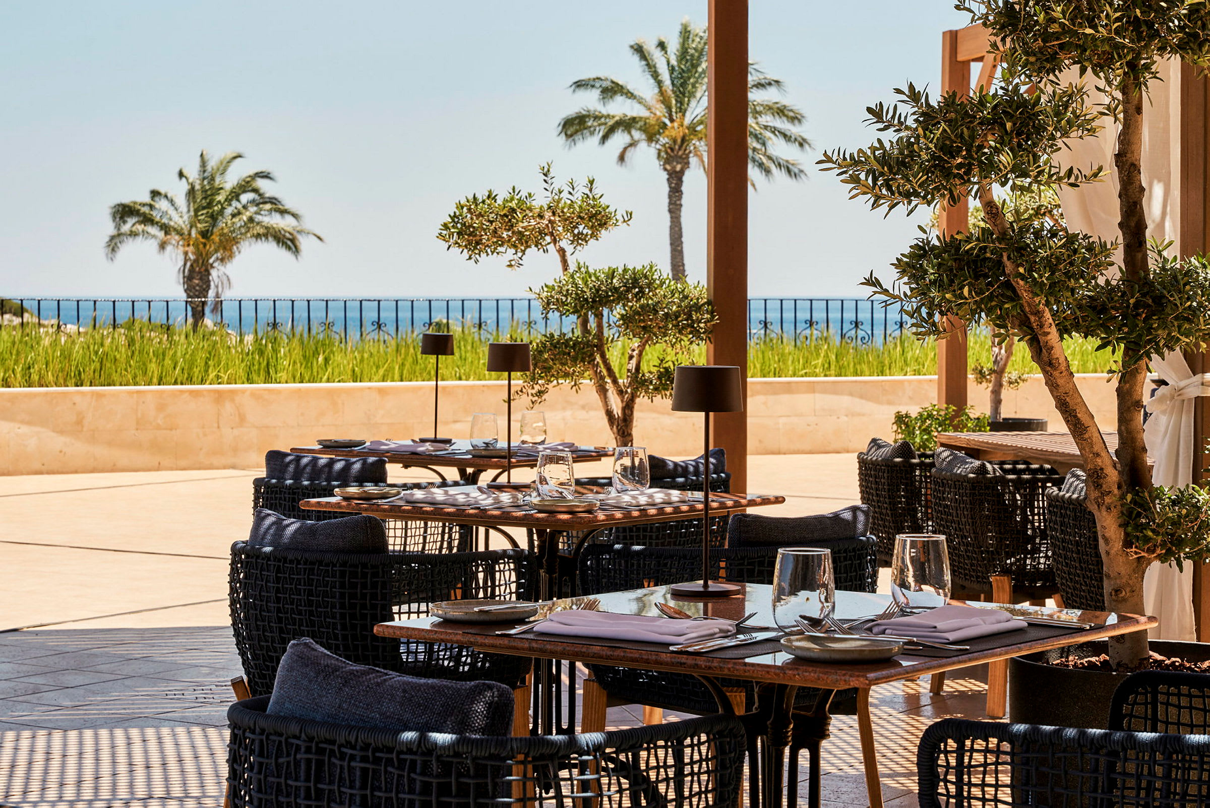 En utomhusrestaurang med dukade bord, omgiven av träd och med utsikt över havet och palmer. (AI-genererad text)