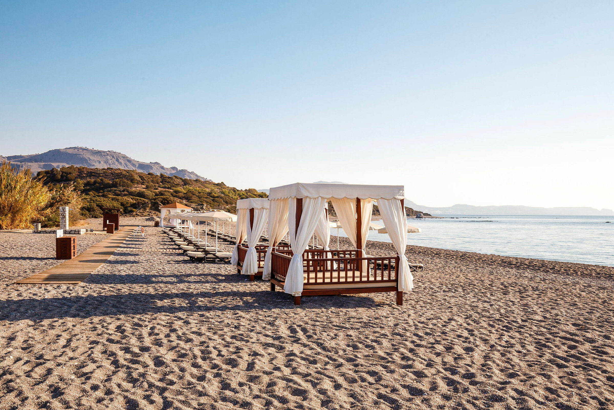 Solstolar och baldakiner på en sandstrand med hav och berg i bakgrunden. (AI-genererad text)