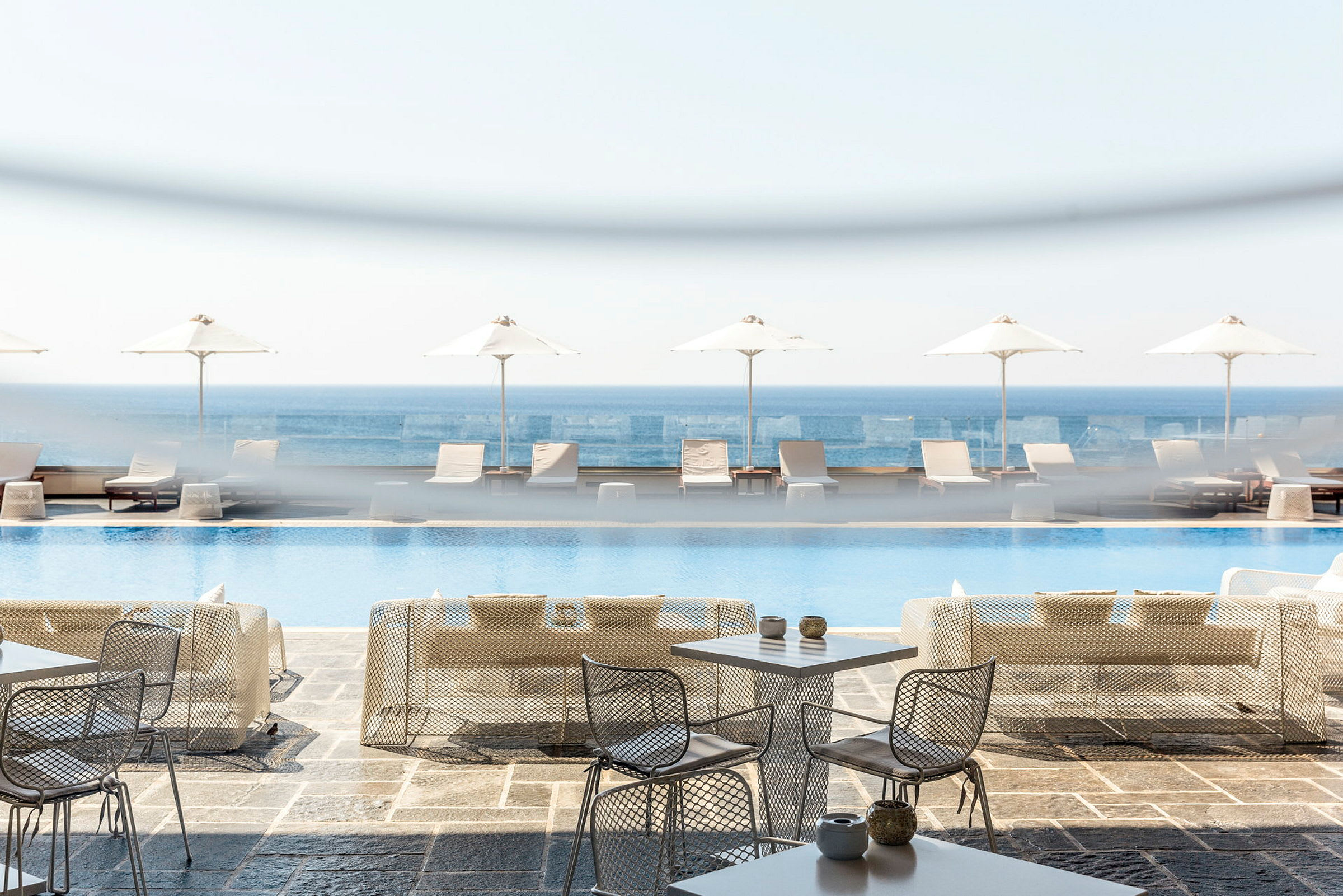 Utomhusbord och stolar framför en pool, solstolar och parasoller med havet i bakgrunden. (AI-genererad text)