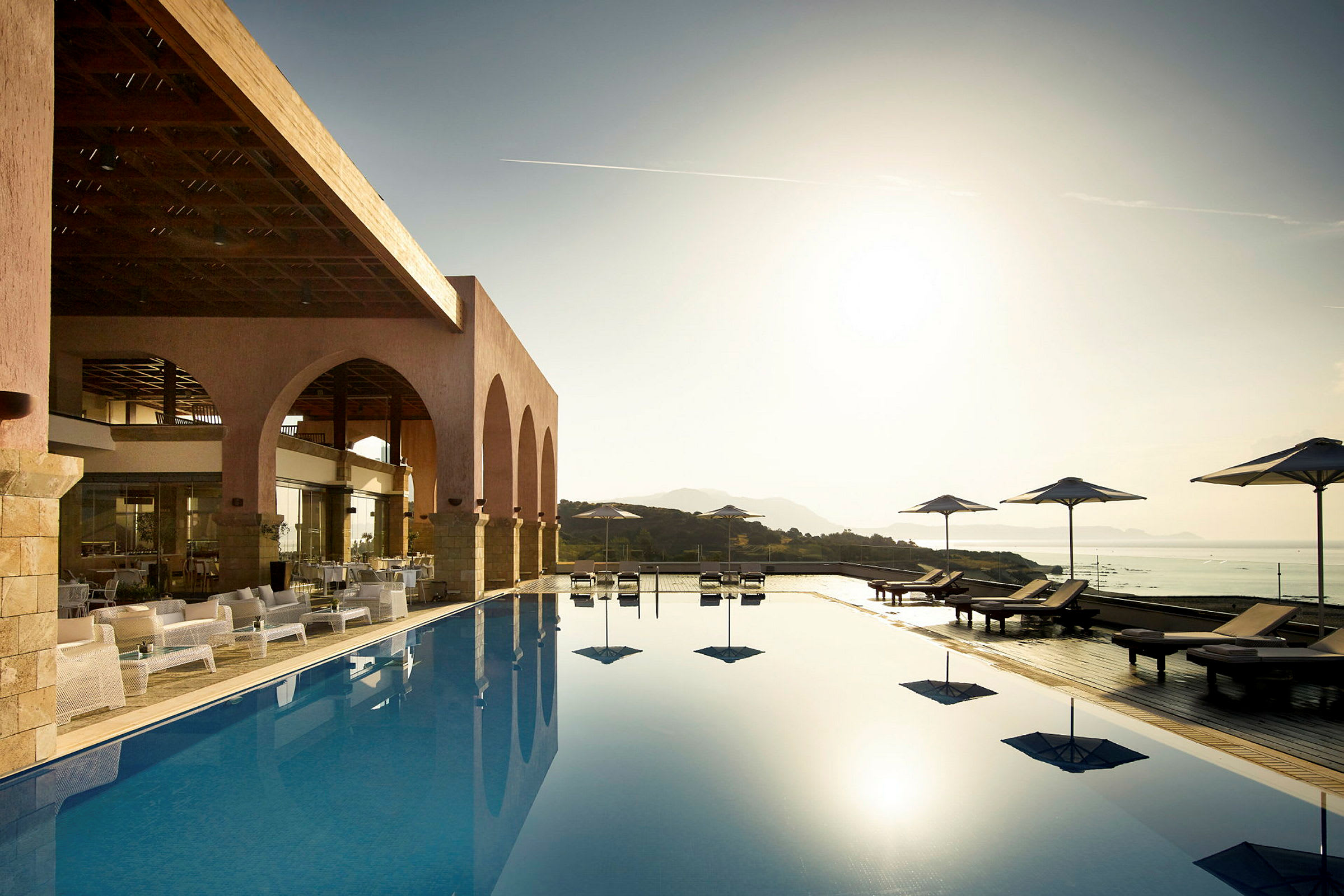 En pool med solstolar och parasoller nära en terrass med välvda öppningar. Solen går ner över landskapet. (AI-genererad text)