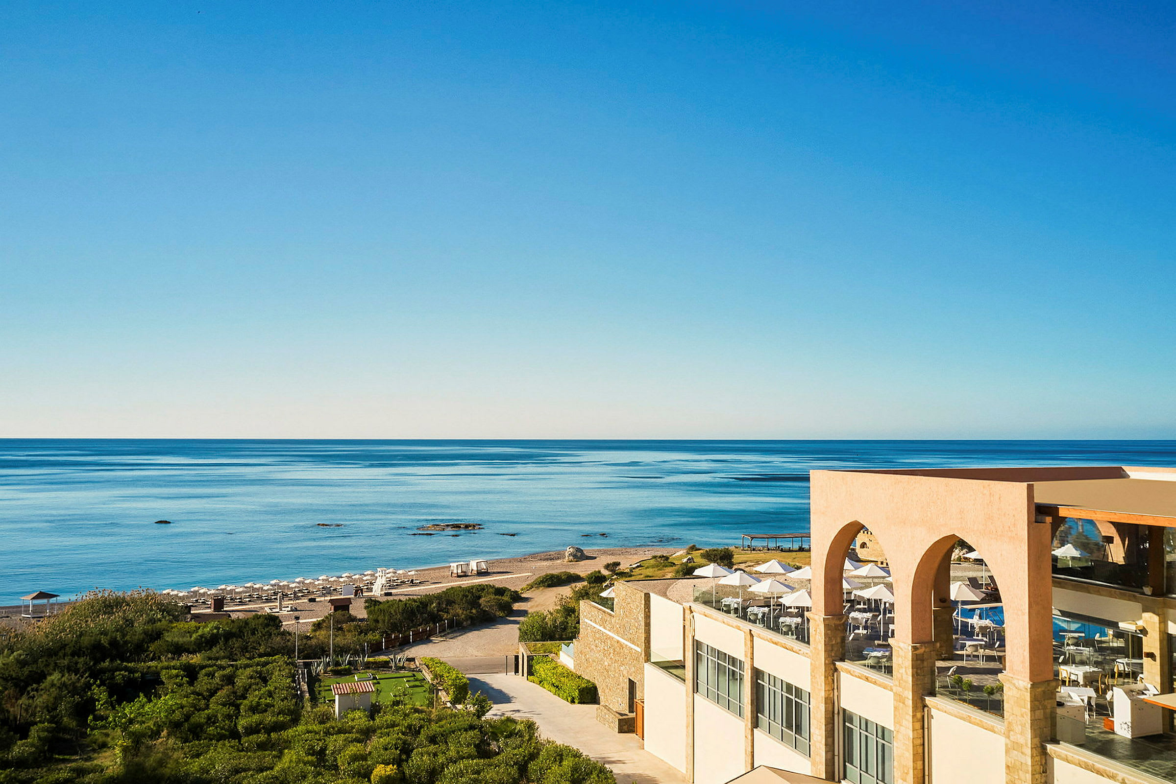 Vy över en sandstrand, havet och ett hotell med terrass i förgrunden. (AI-genererad text)