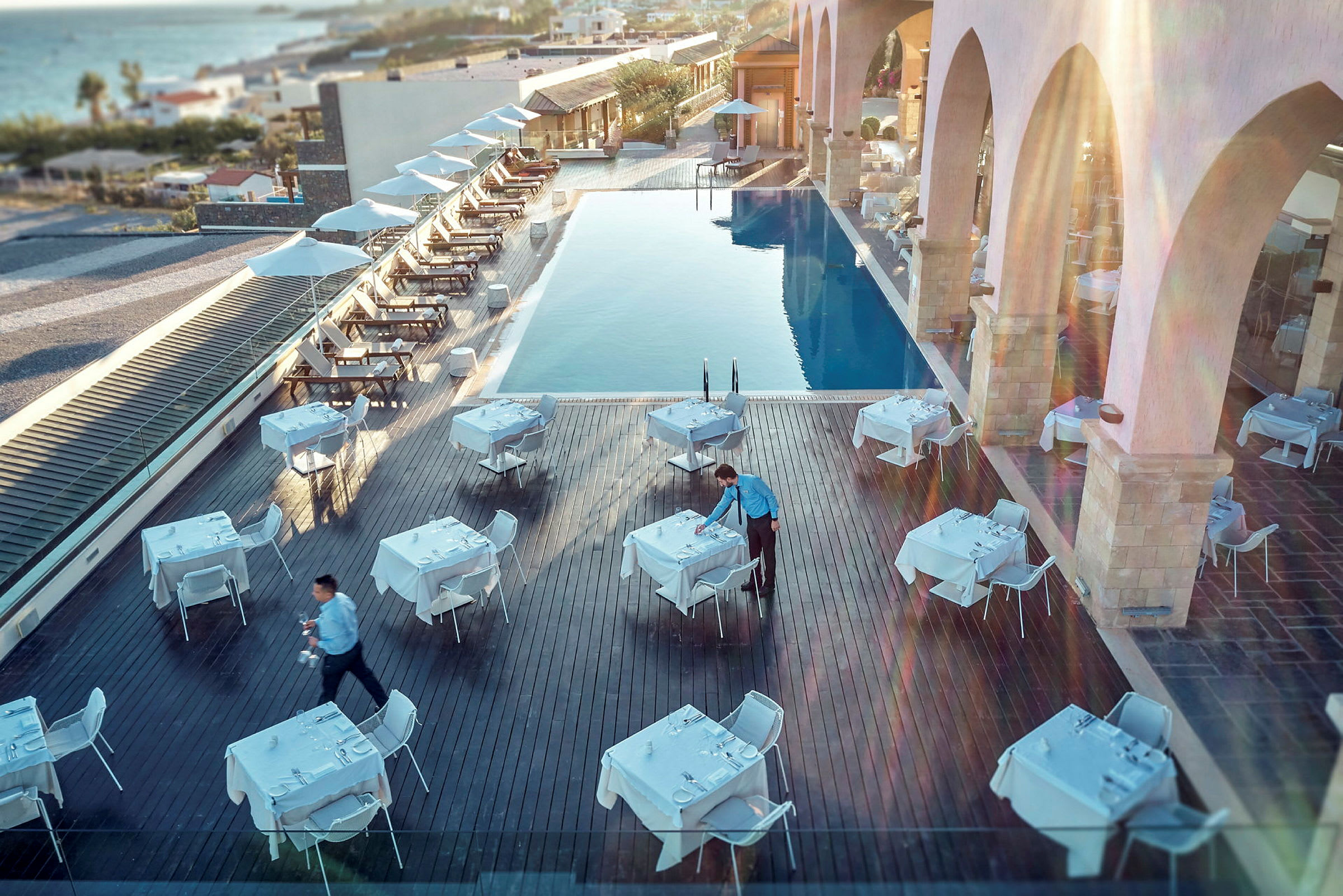 Utomhusrestaurang vid en pool med uppställda bord och stolar, servitör förbereder dukning. (AI-genererad text)