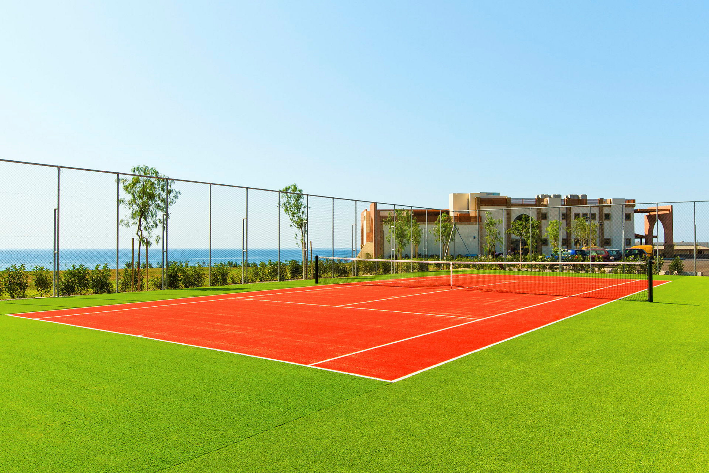 En tennisplan med röd beläggning omgiven av grönt gräs, med havet och en byggnad i bakgrunden. (AI-genererad text)