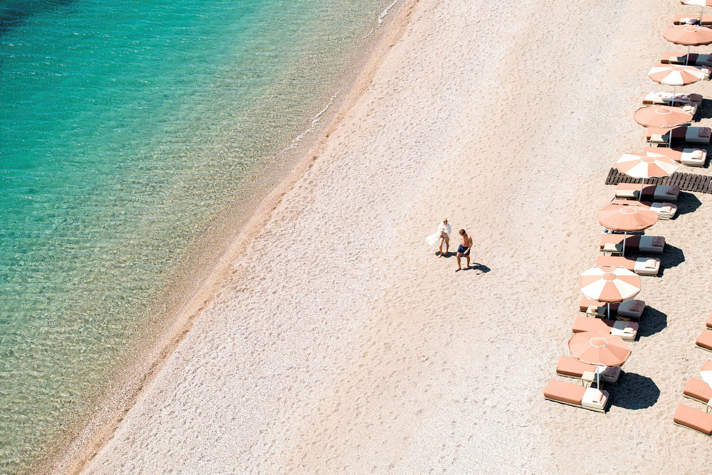 En strand med klart vatten och solparasoller längs ena sidan. Två personer promenerar på sanden. (AI-genererad text)