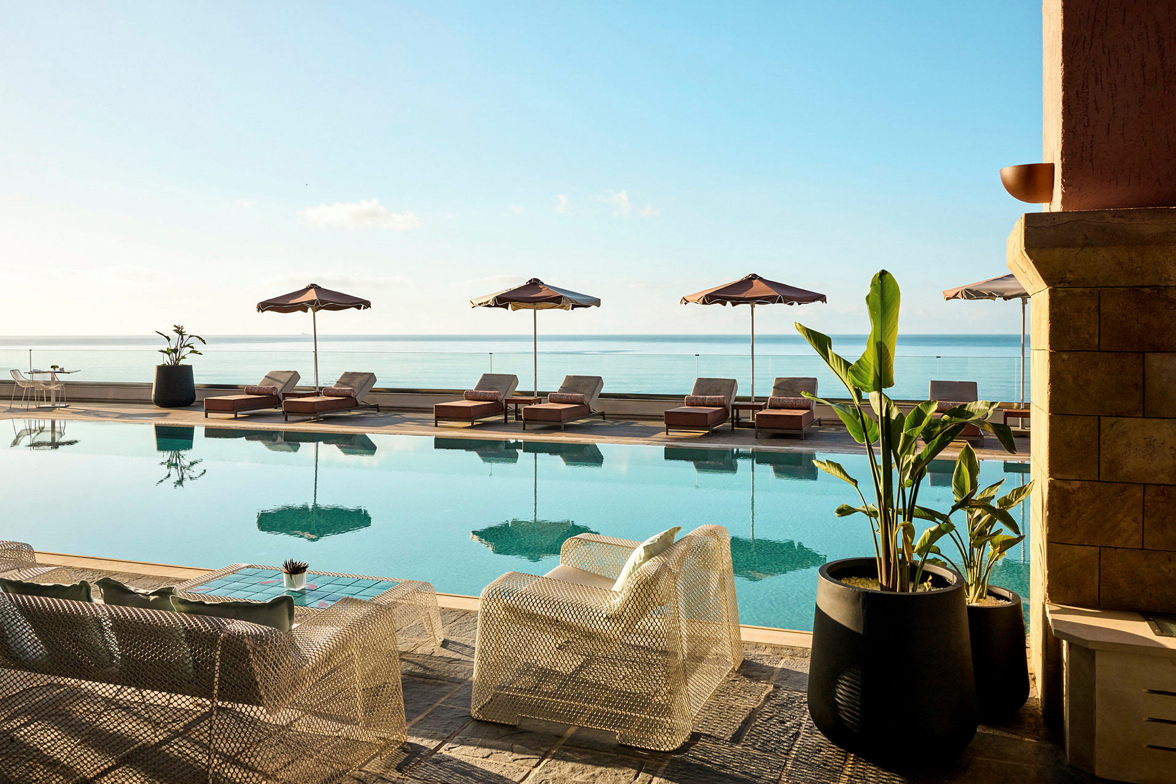 En swimmingpool med solstolar och parasoller vid sidan och utsikt över havet. Framför poolen står stolar och bord samt stora krukväxter. (AI-genererad text)