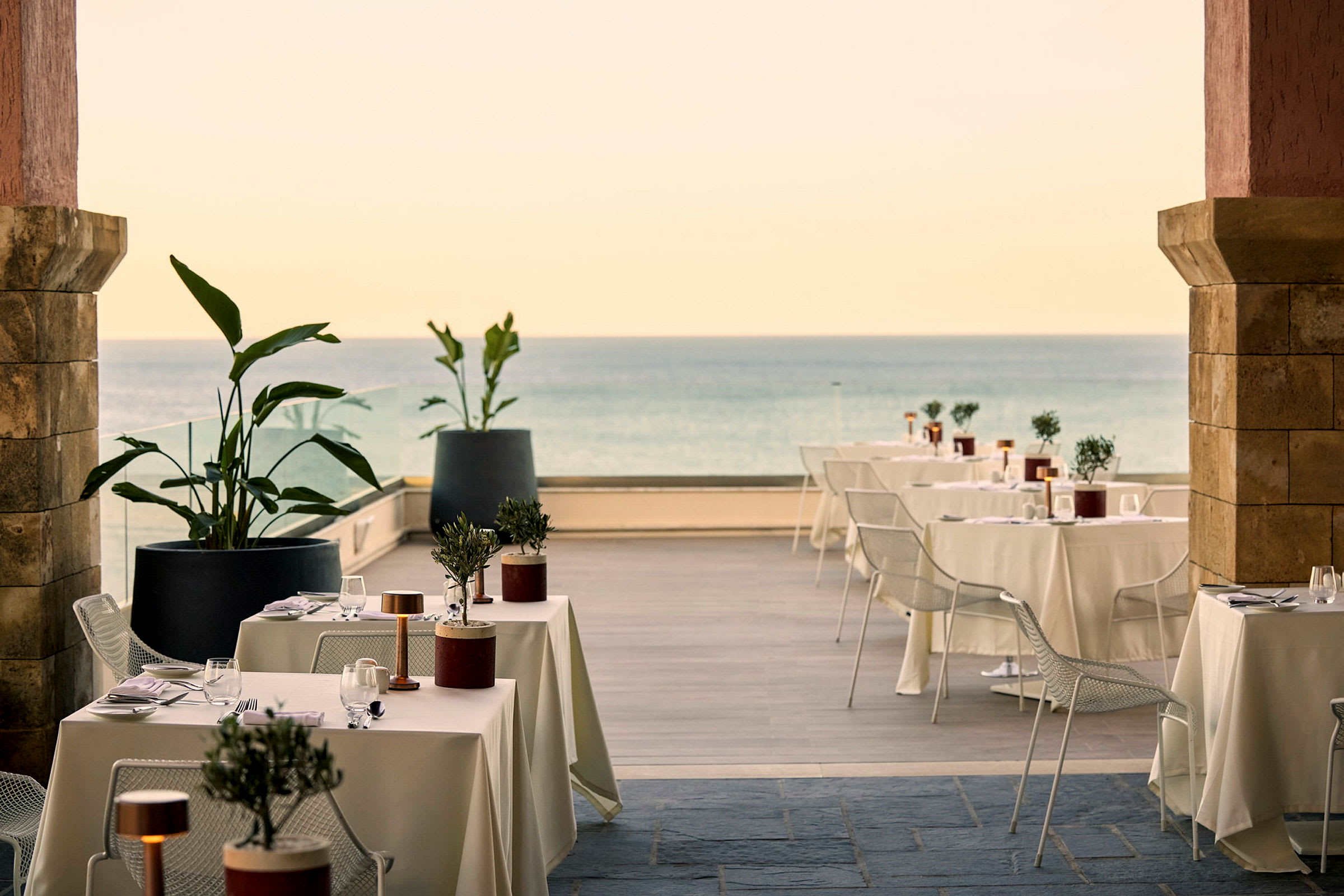 Utomhusrestaurang med dukade bord, stolar och dekorativa växter nära havet. (AI-genererad text)