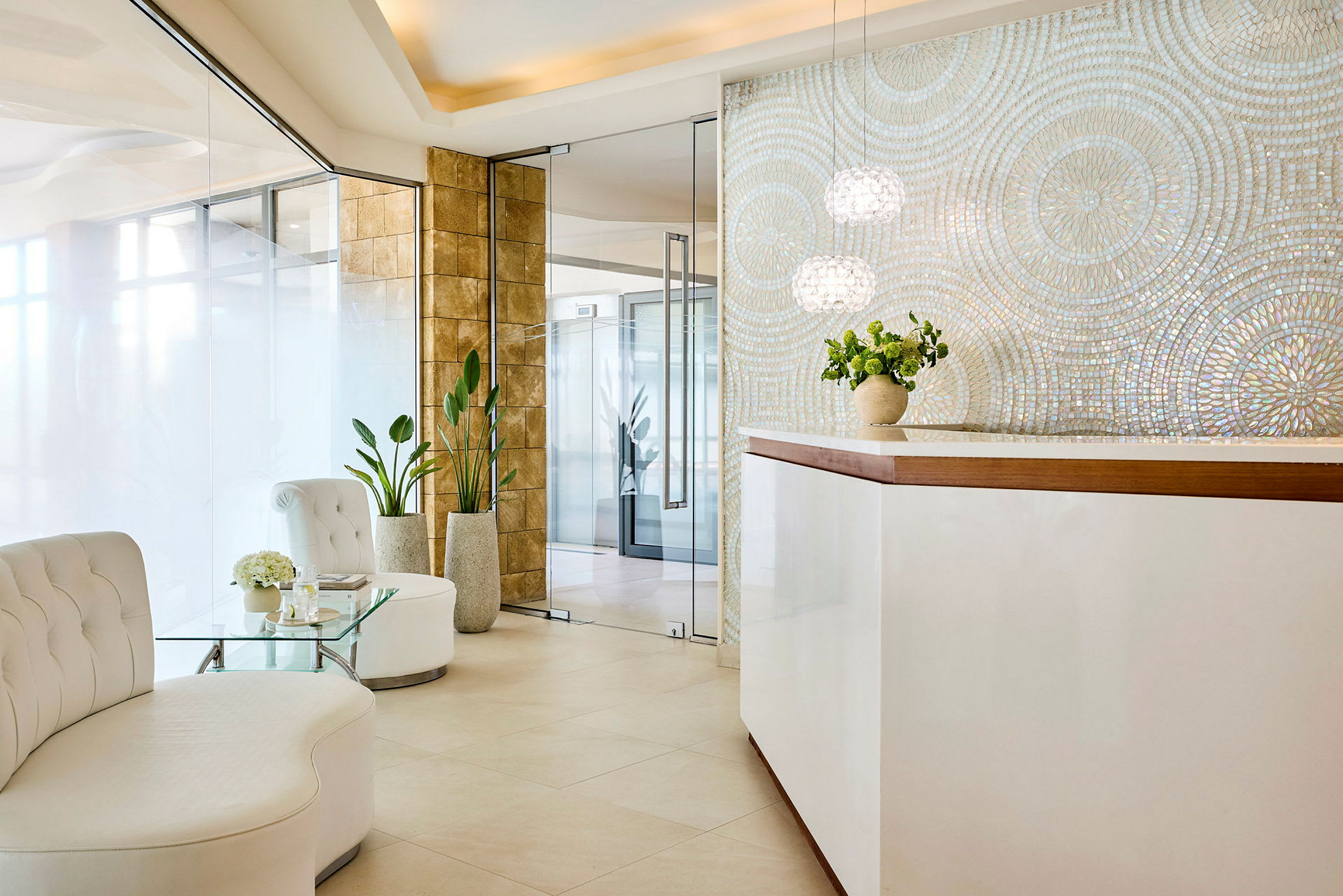 En modern receptionsområde med vita möbler, glasbord och grönska. En stilren vägg med iriserande mosaik finns bakom disken. (AI-genererad text)