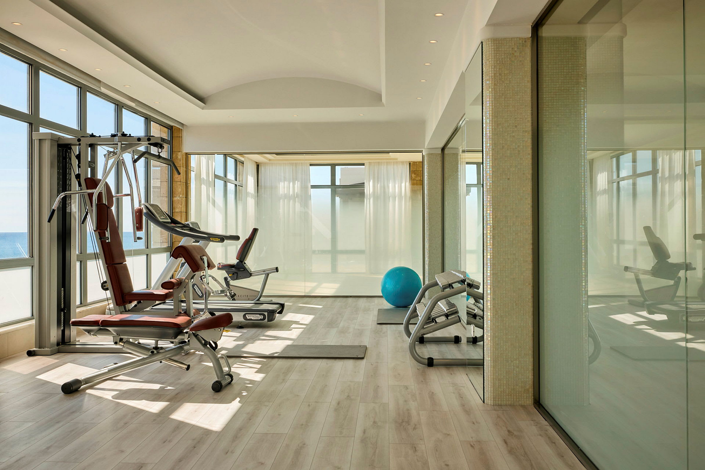 Ett gym med träningsmaskiner, en blå träningsboll och stora fönster som släpper in naturligt ljus. (AI-genererad text)
